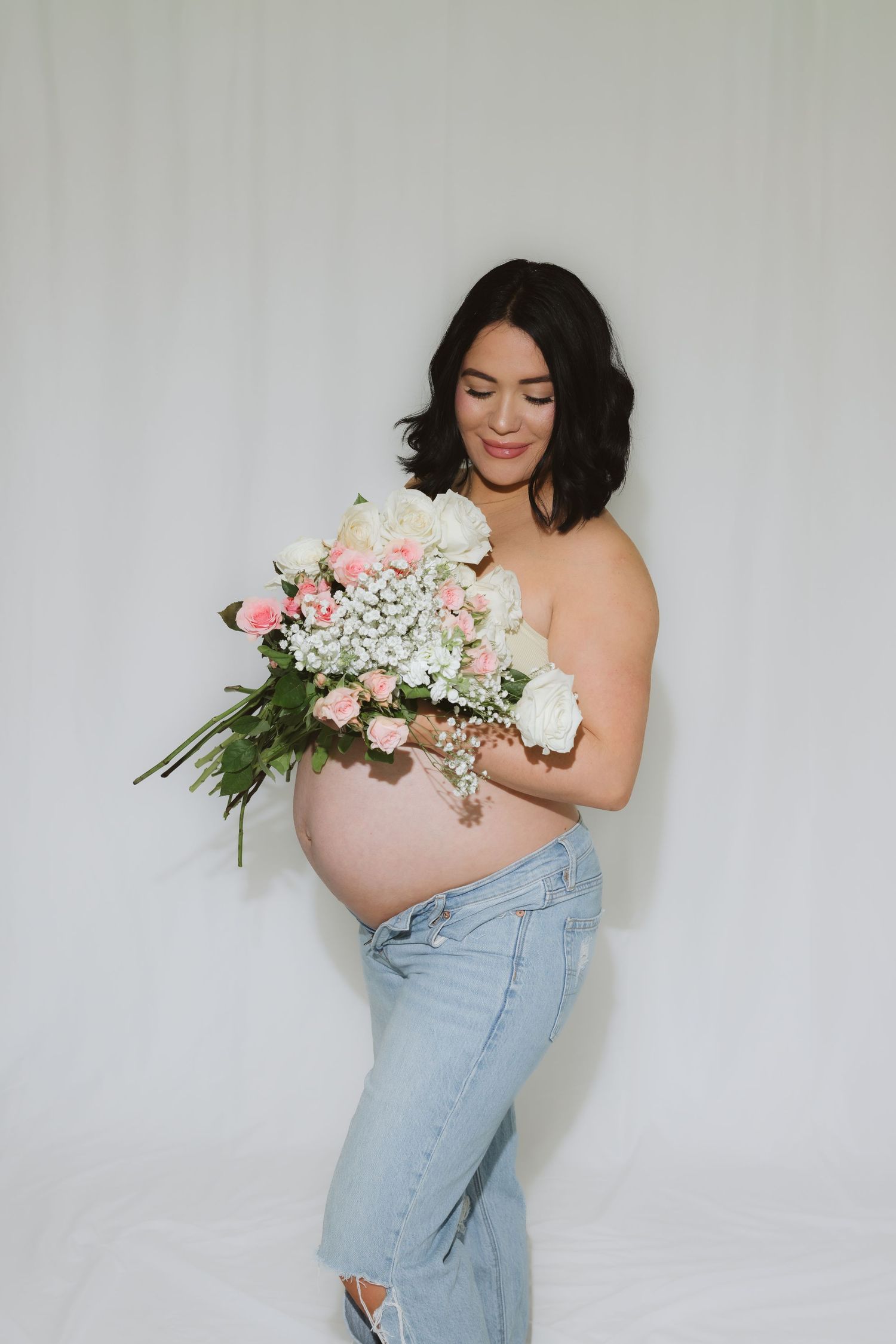 Pregnant woman holding pink and white flower bouquet, wearing ripped jeans, posing against white backdrop.