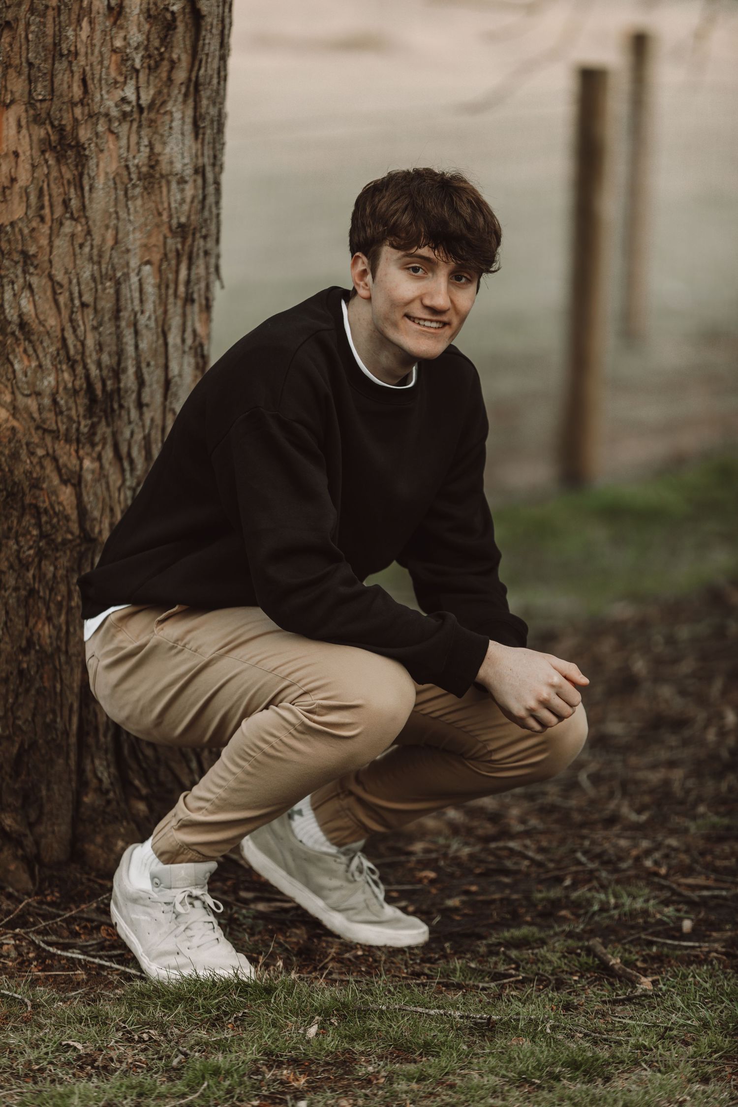 Person in black sweater and khaki pants crouching near tree trunk wearing white sneakers.