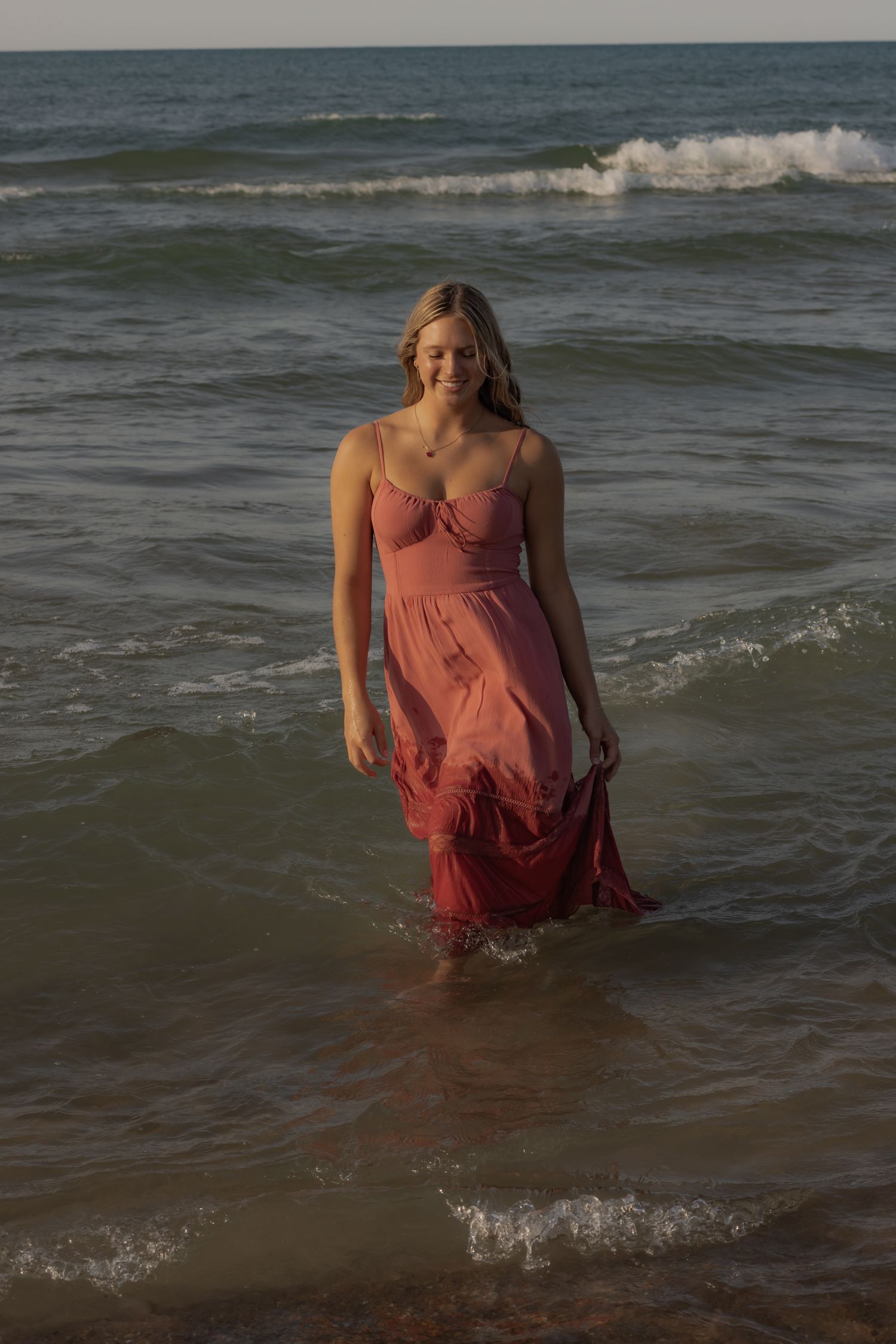 Figure in coral dress standing in ocean waves at sunset.
