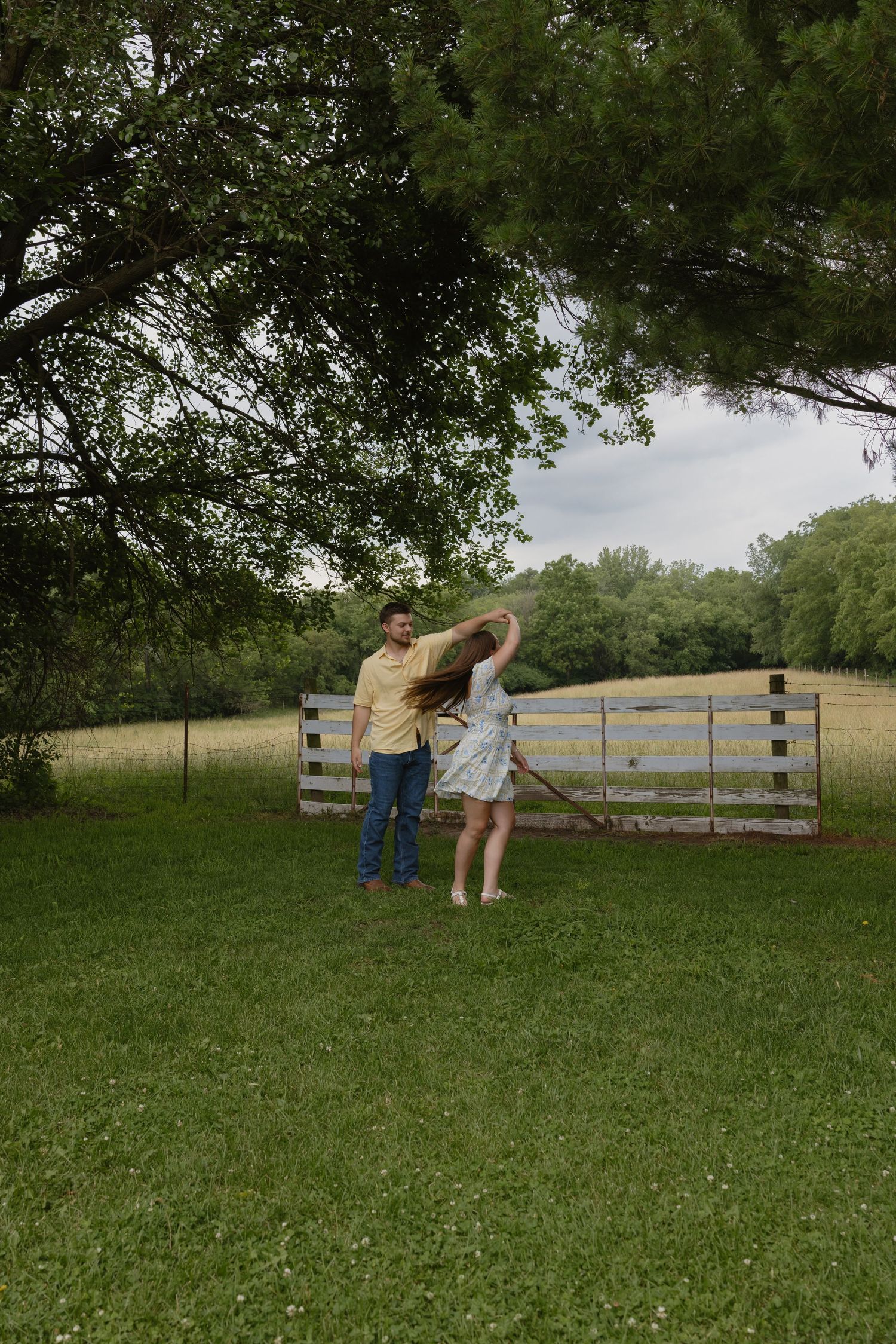 Couple dancing together on green grass near a wooden fence in a scenic rural countryside setting.