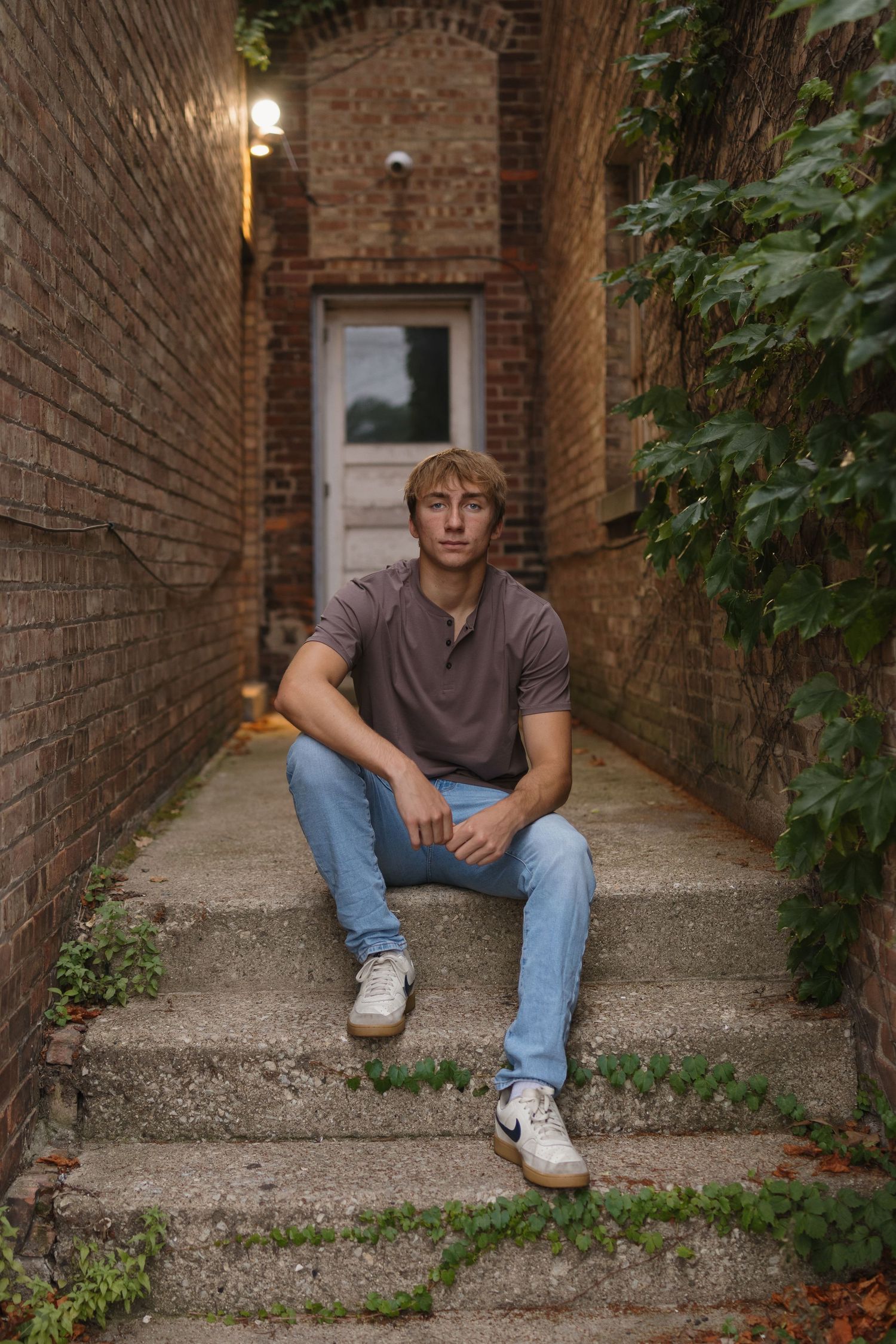 Young man in blue jeans and brown polo sits on brick alley steps surrounded by ivy walls.