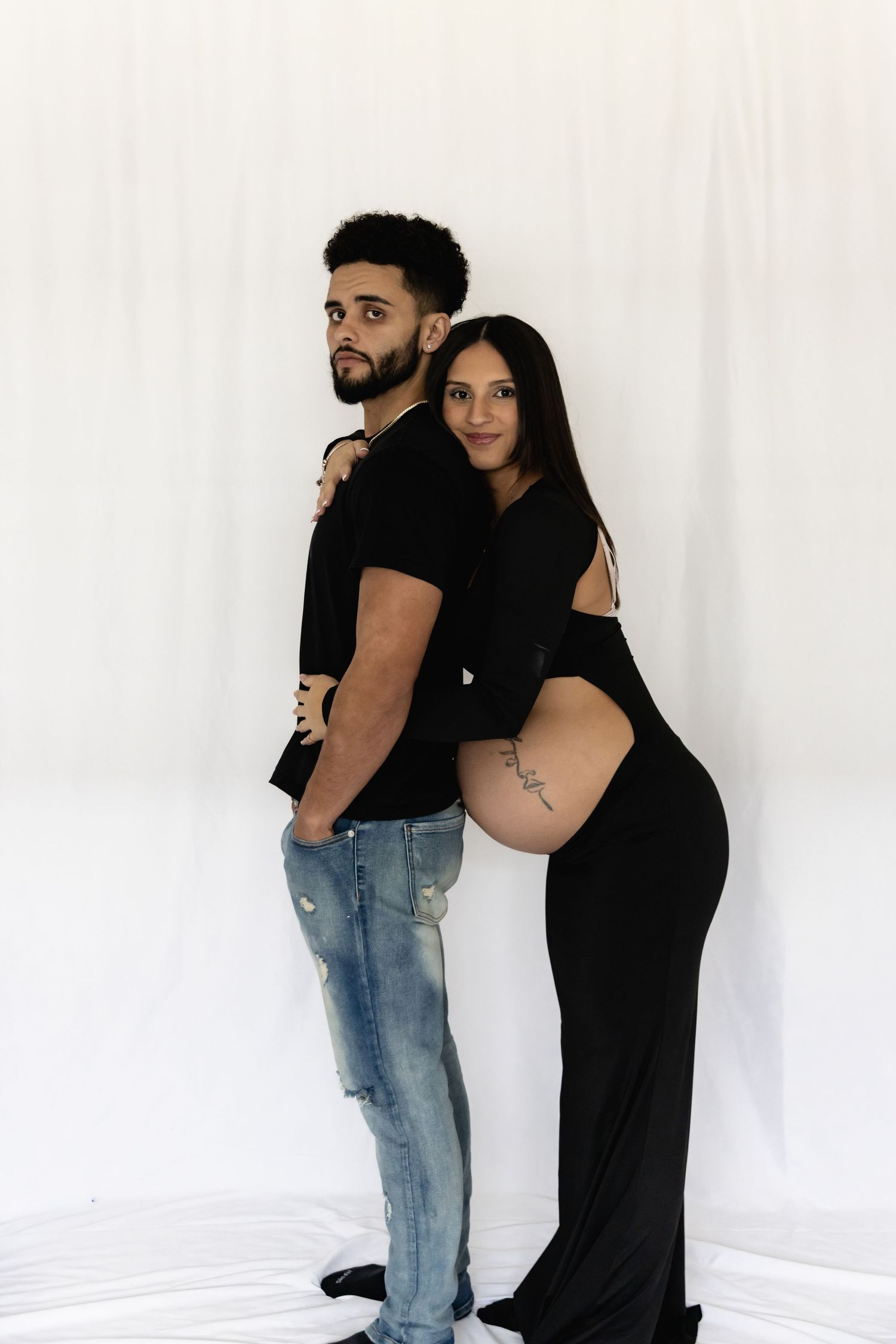 A couple in black clothing poses together against a white backdrop during a maternity photoshoot.