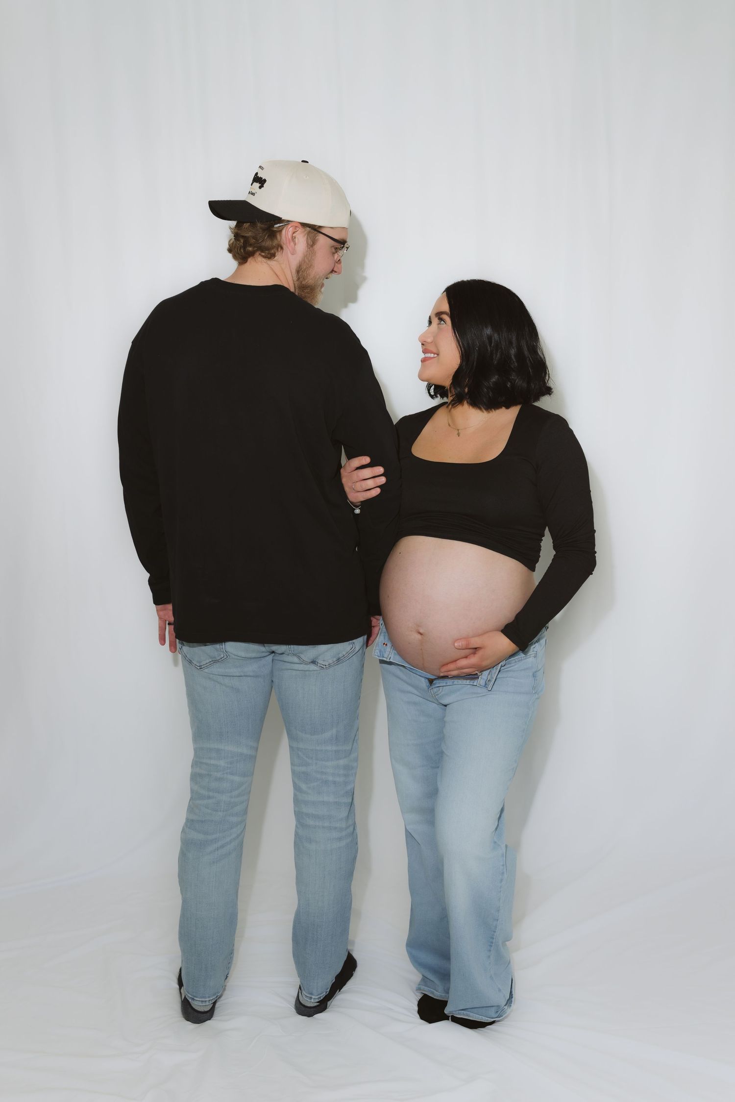 Pregnant couple in matching black tops and jeans posing together, woman cradling baby bump, man wearing cream cap.