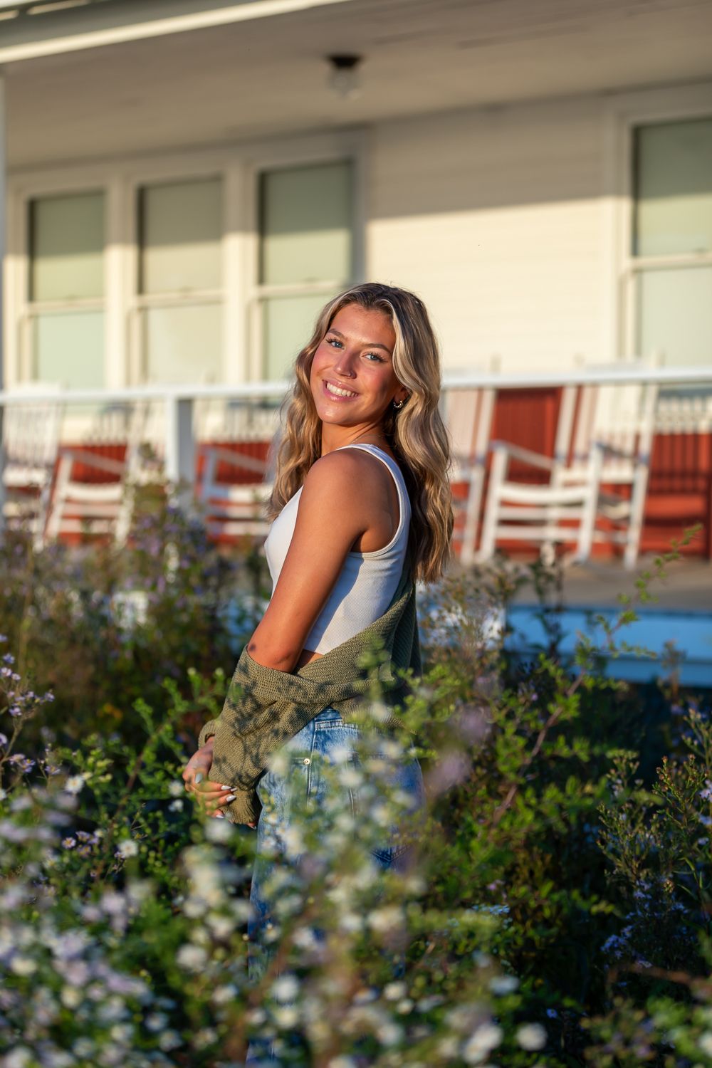 Smiling blonde woman in white tank and green jacket standing among wildflowers outside a building with red rocking chairs.