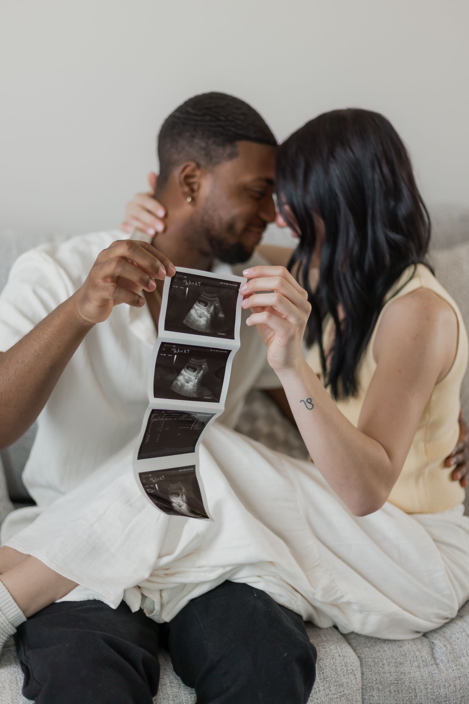Happy couple sitting on couch holding ultrasound photos, touching foreheads in an intimate pregnancy announcement moment.