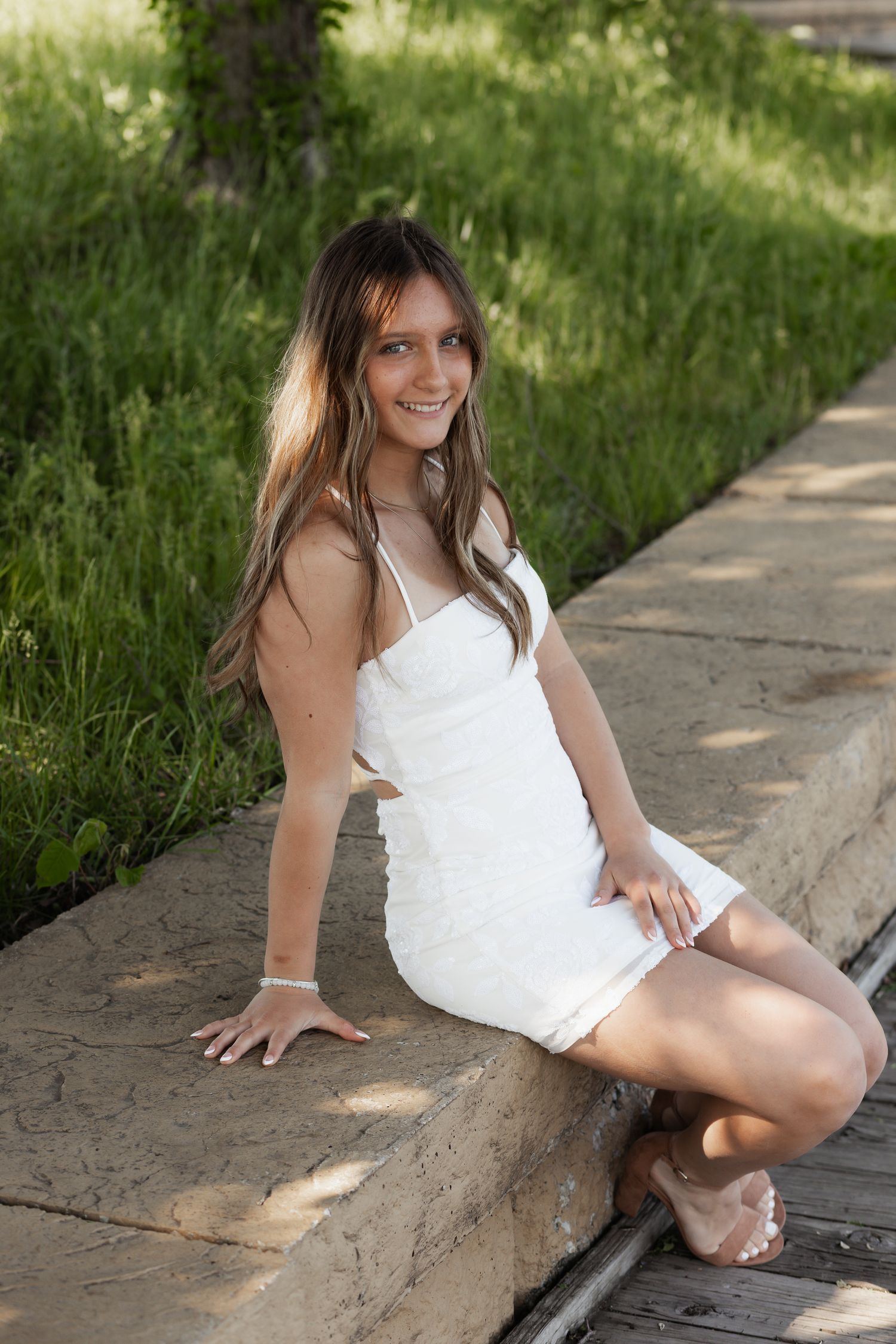 A smiling young woman in a white mini dress sitting outdoors on a stone ledge surrounded by greenery.