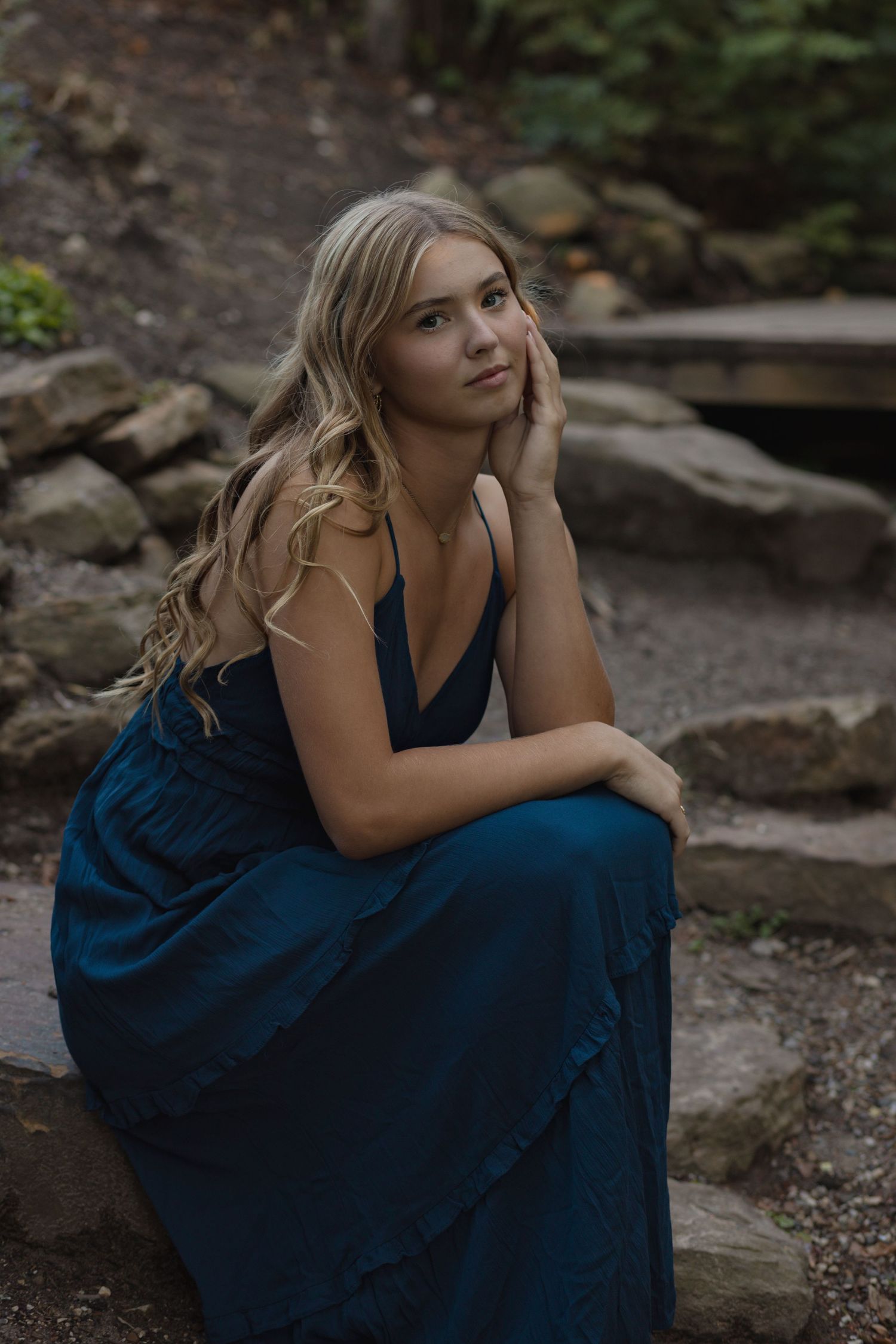 Woman in navy blue dress sitting thoughtfully on rocky surface in natural setting.