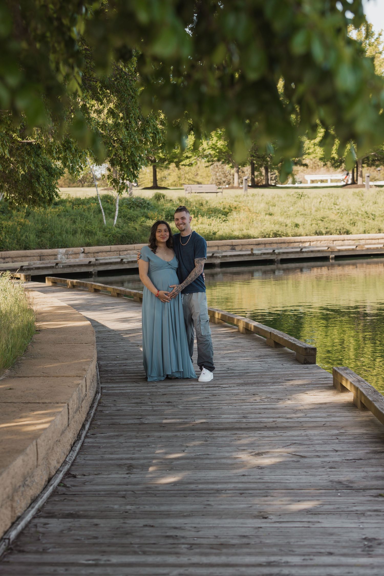 Couple posing on wooden boardwalk by pond, woman in blue maternity gown, man in casual attire, surrounded by lush greenery.