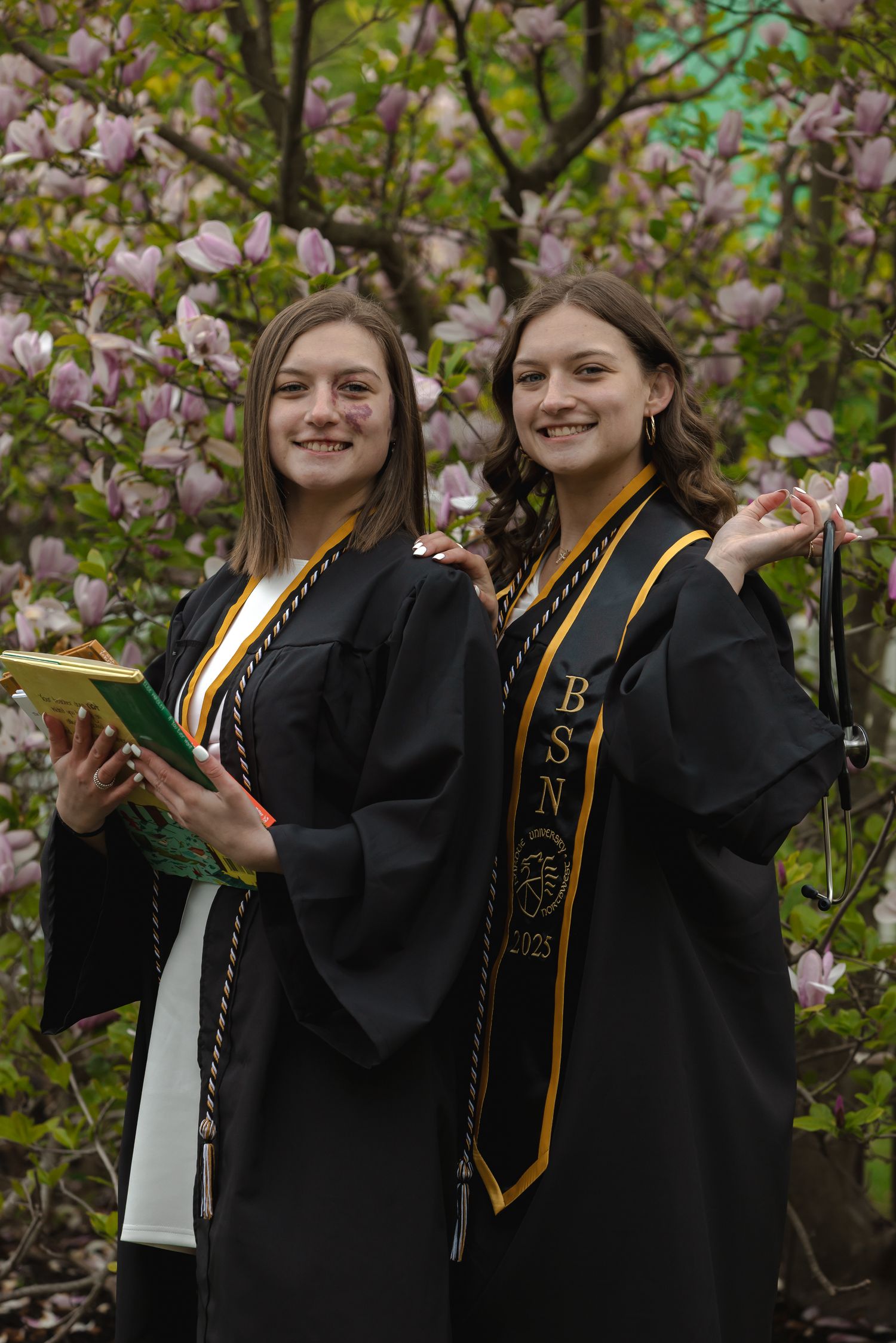 Graduates in black robes and gold stoles celebrate among pink cherry blossoms during spring commencement.