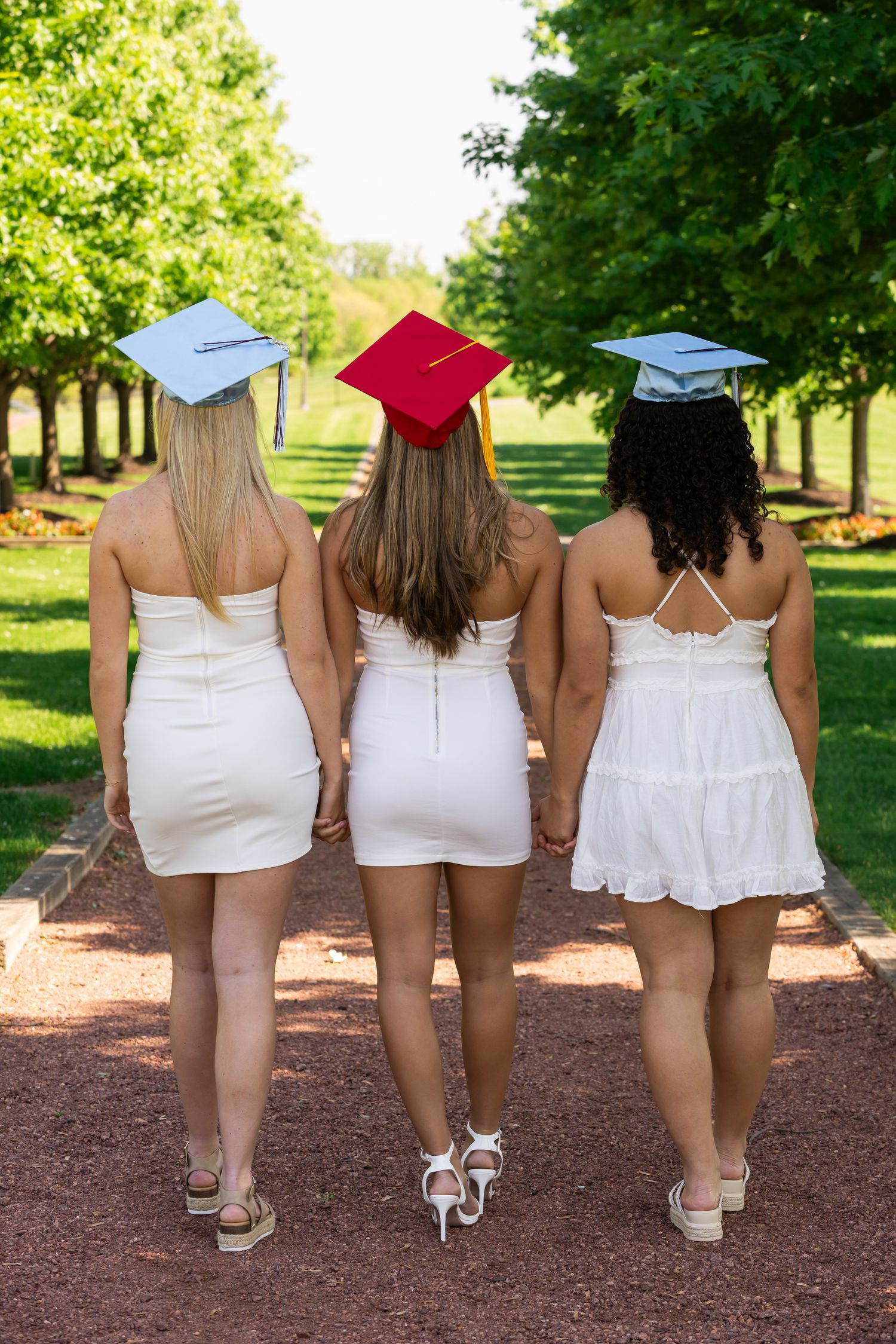 Back view of three graduates in white dresses and colorful caps walking on mulch path.