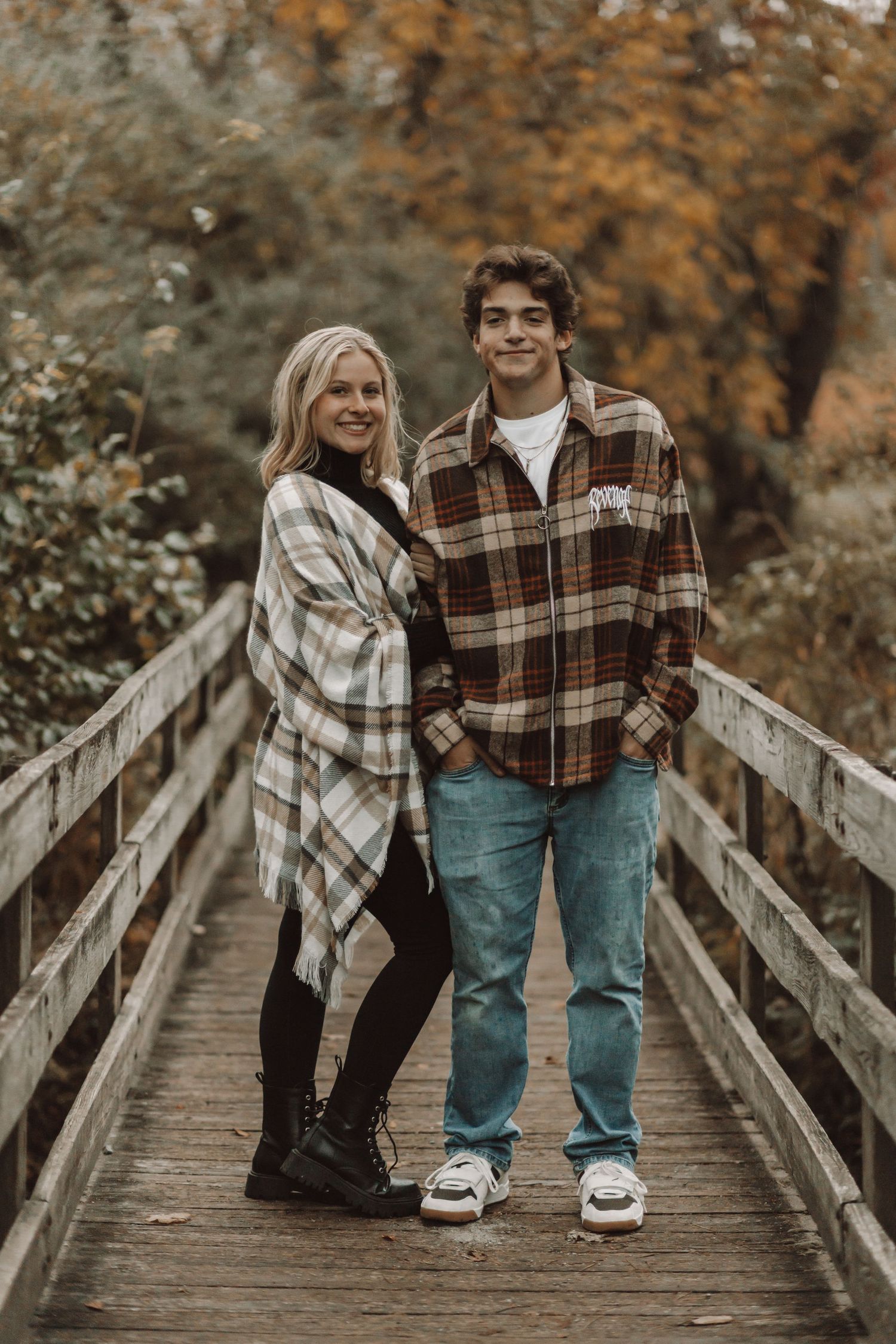 Young couple in plaid shirts smiling on a wooden bridge surrounded by autumn foliage.