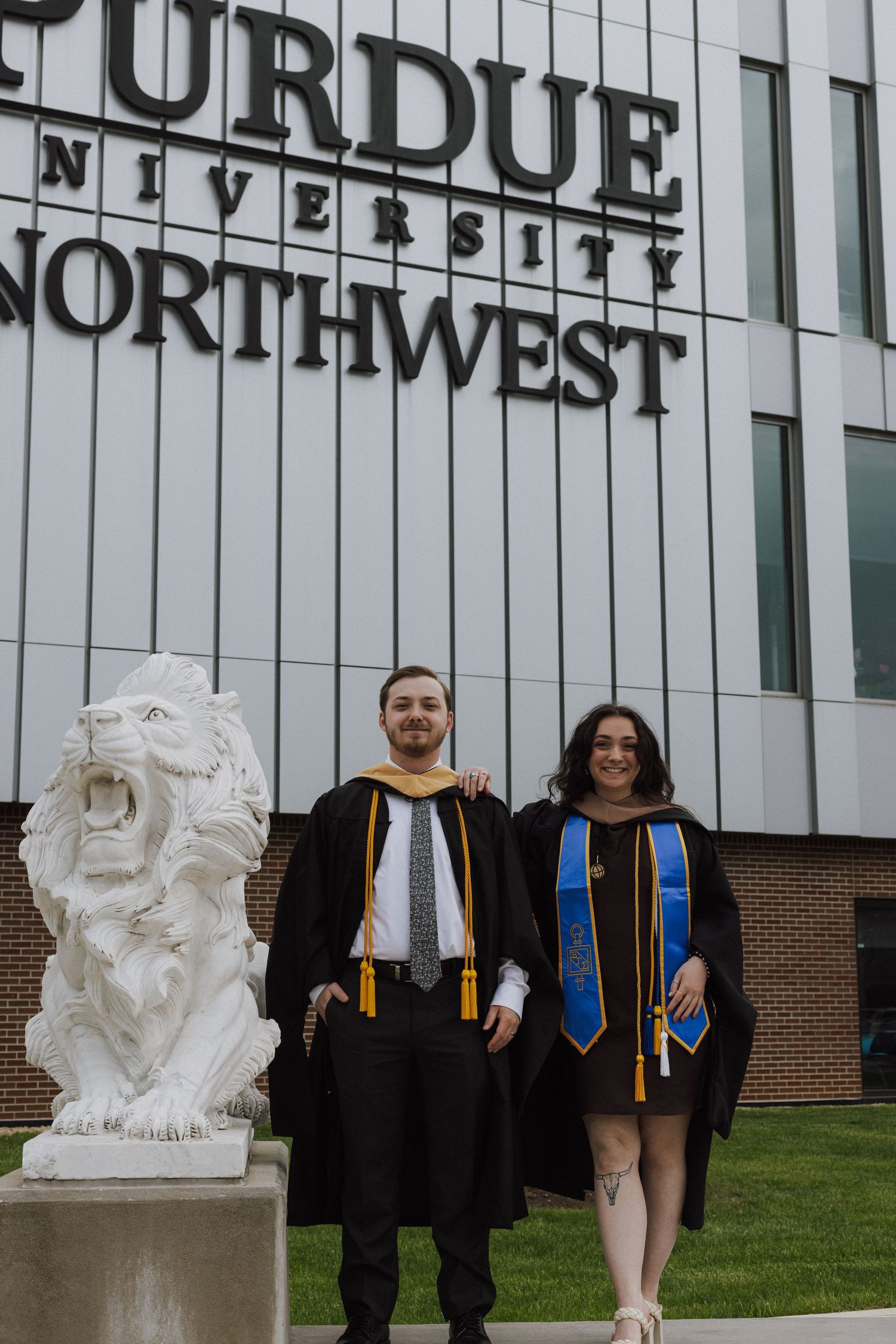 Two graduates in black robes pose proudly before Purdue University Northwest sign beside a lion statue.