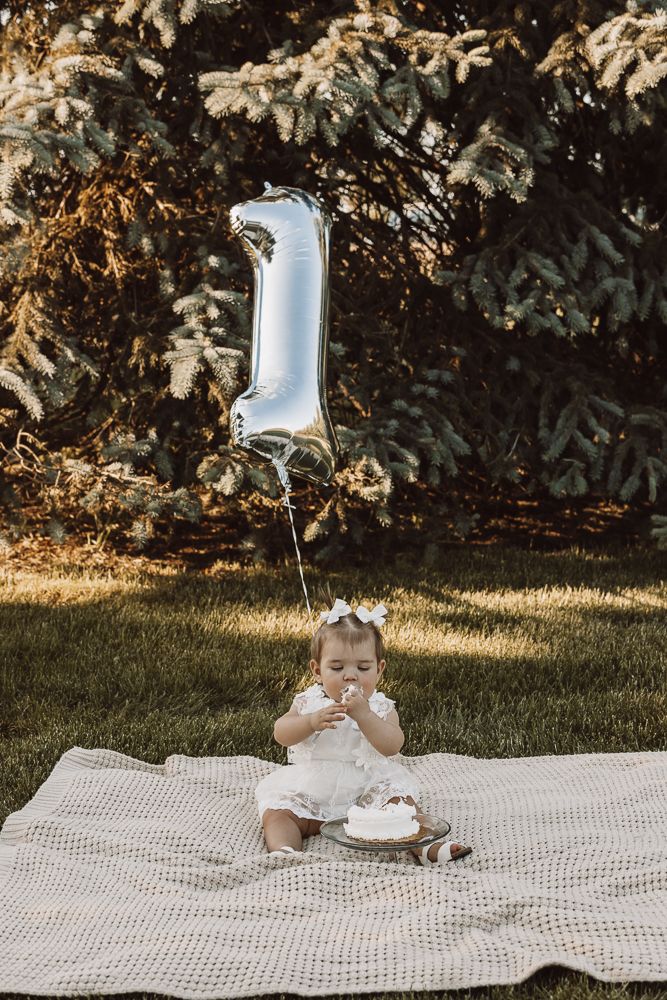 Baby sitting on white blanket in garden having snack time outdoors.
