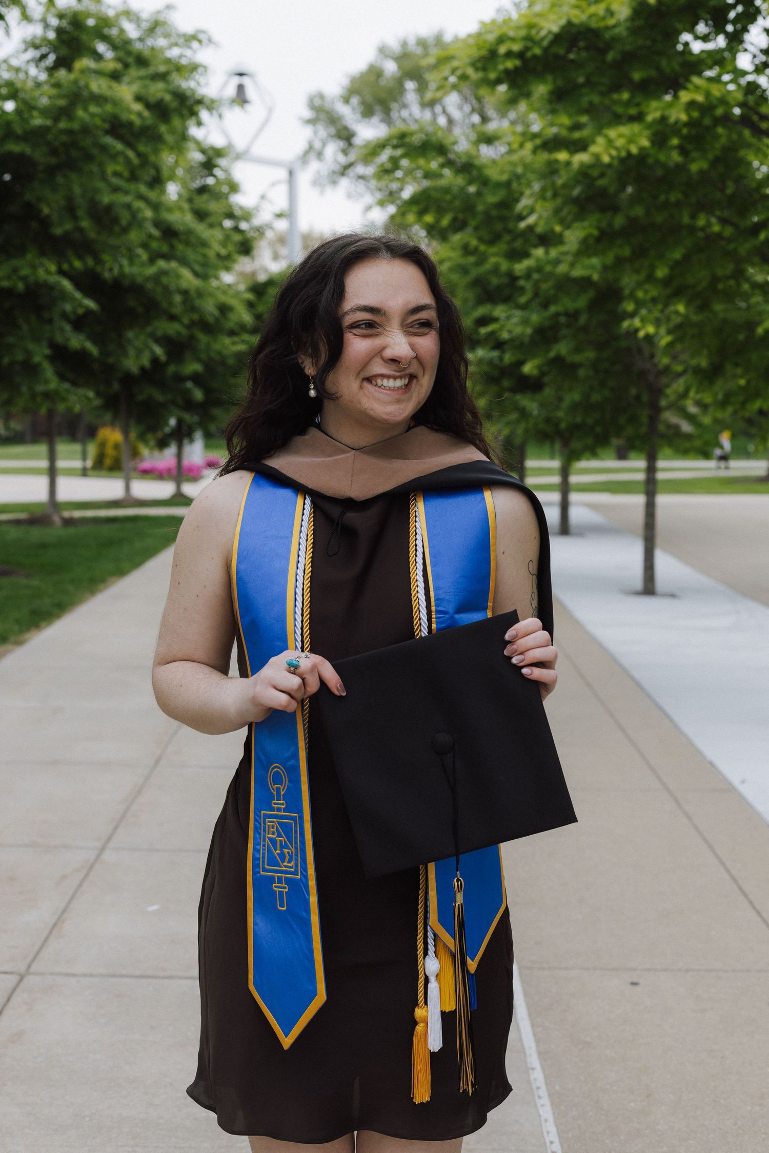 Smiling college graduate in black gown and blue stole holds cap outdoors on campus walkway surrounded by green trees.