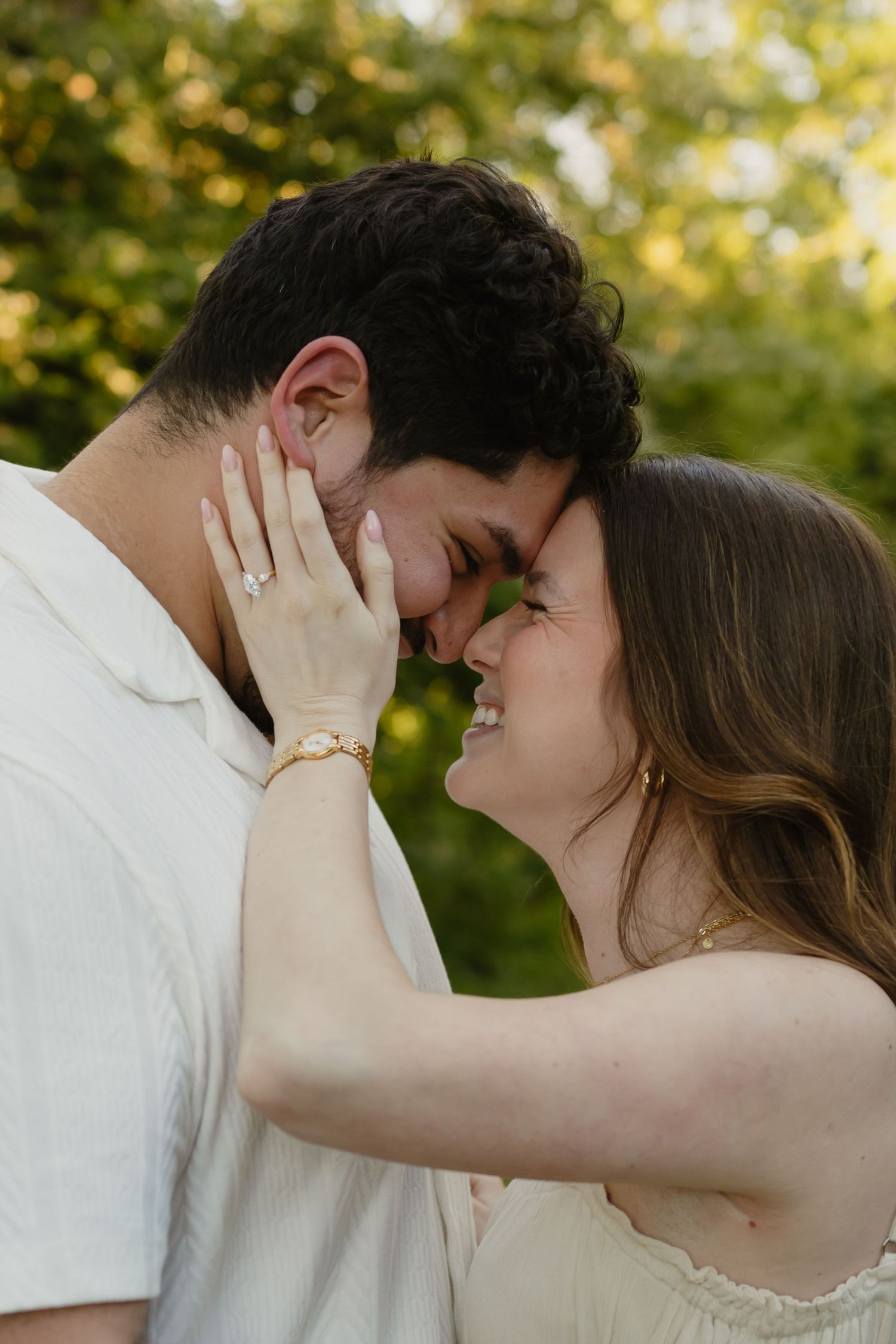 Couple sharing a romantic moment outdoors, woman smiling with engagement ring visible, green trees in background.