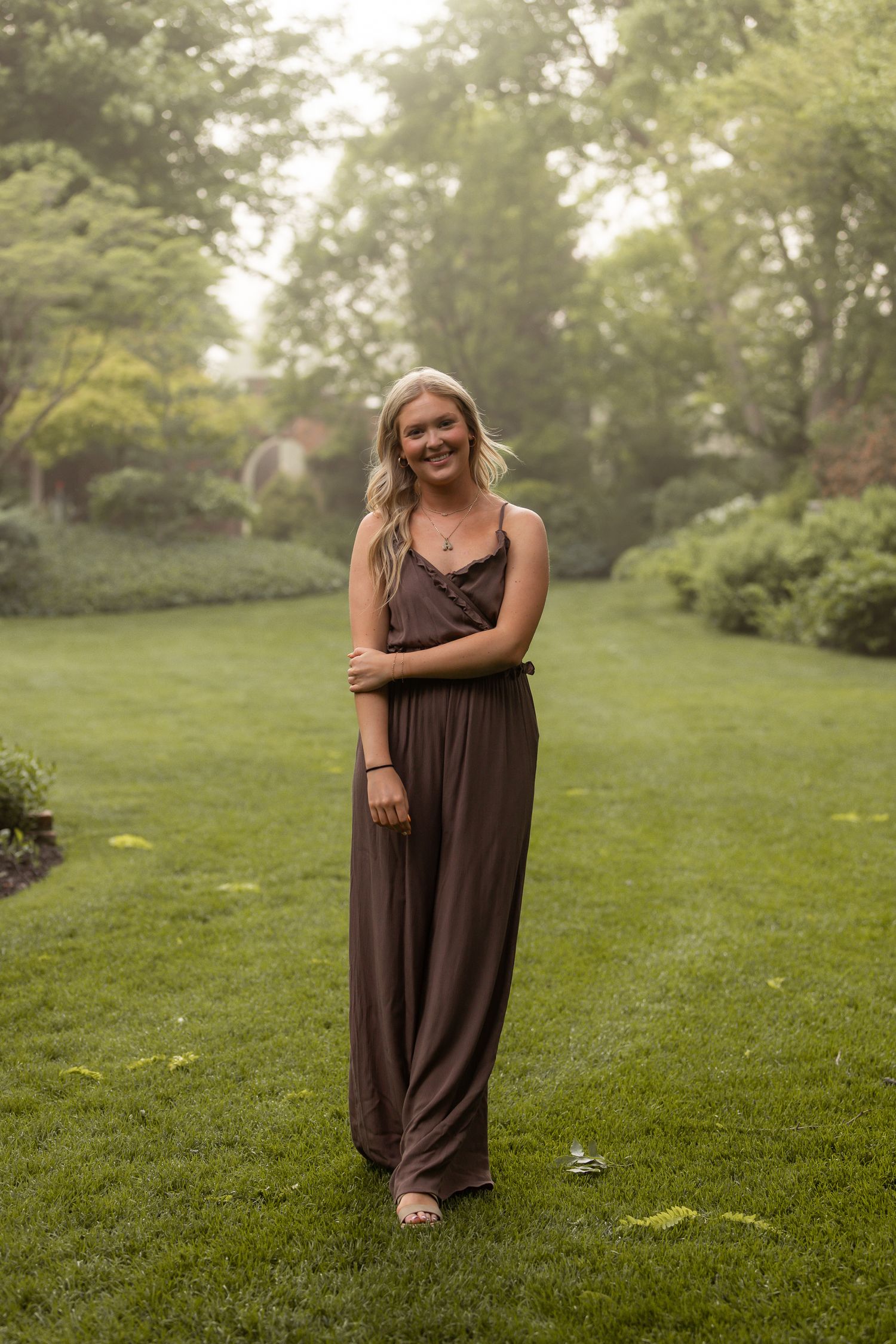 Woman in brown maxi dress poses playfully on green lawn during outdoor photo session.