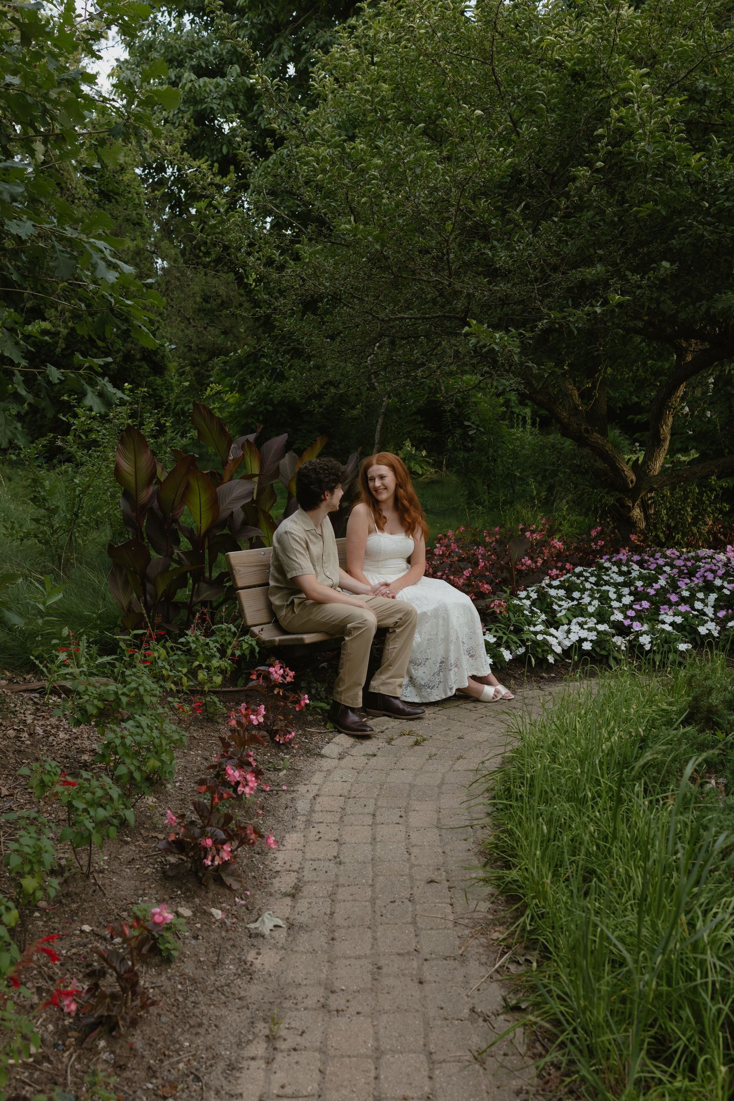 A couple sits on a garden bench surrounded by colorful flowers and lush greenery on a brick pathway.