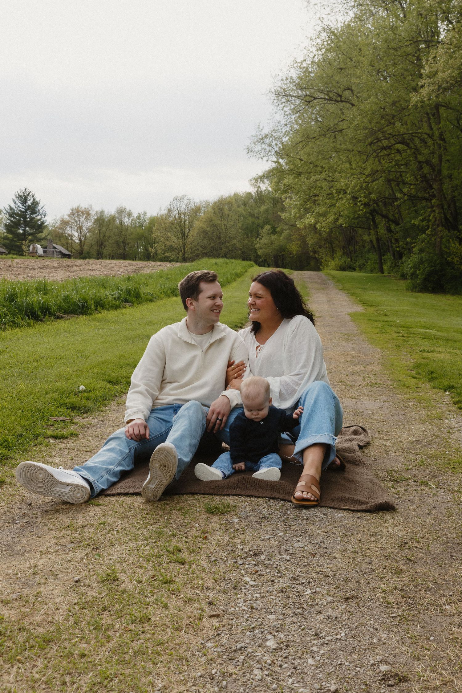 A young family sits on a blanket on a gravel path, smiling at each other with their baby on a cloudy day.