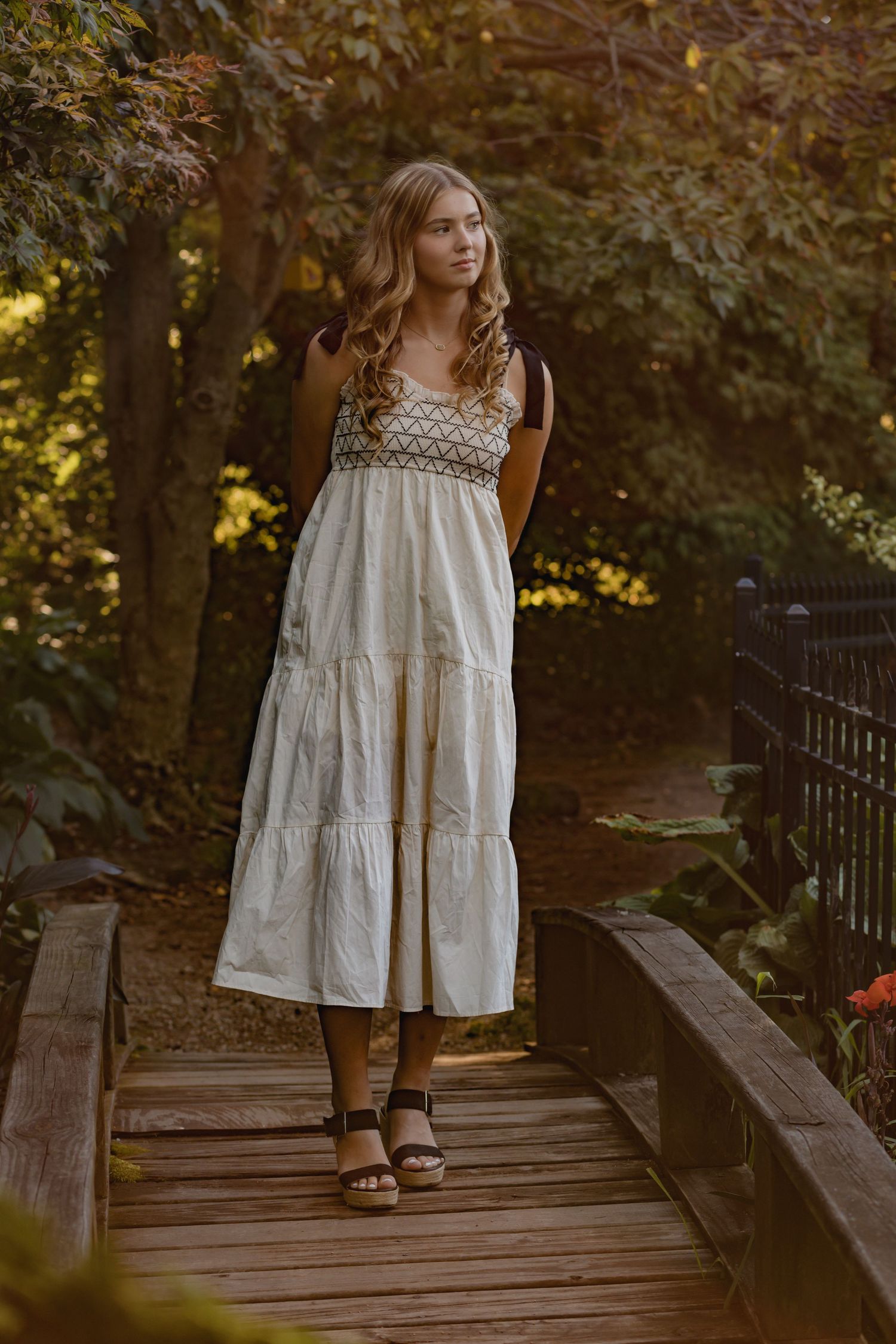 Woman in flowing white summer dress standing on wooden bridge in garden setting.