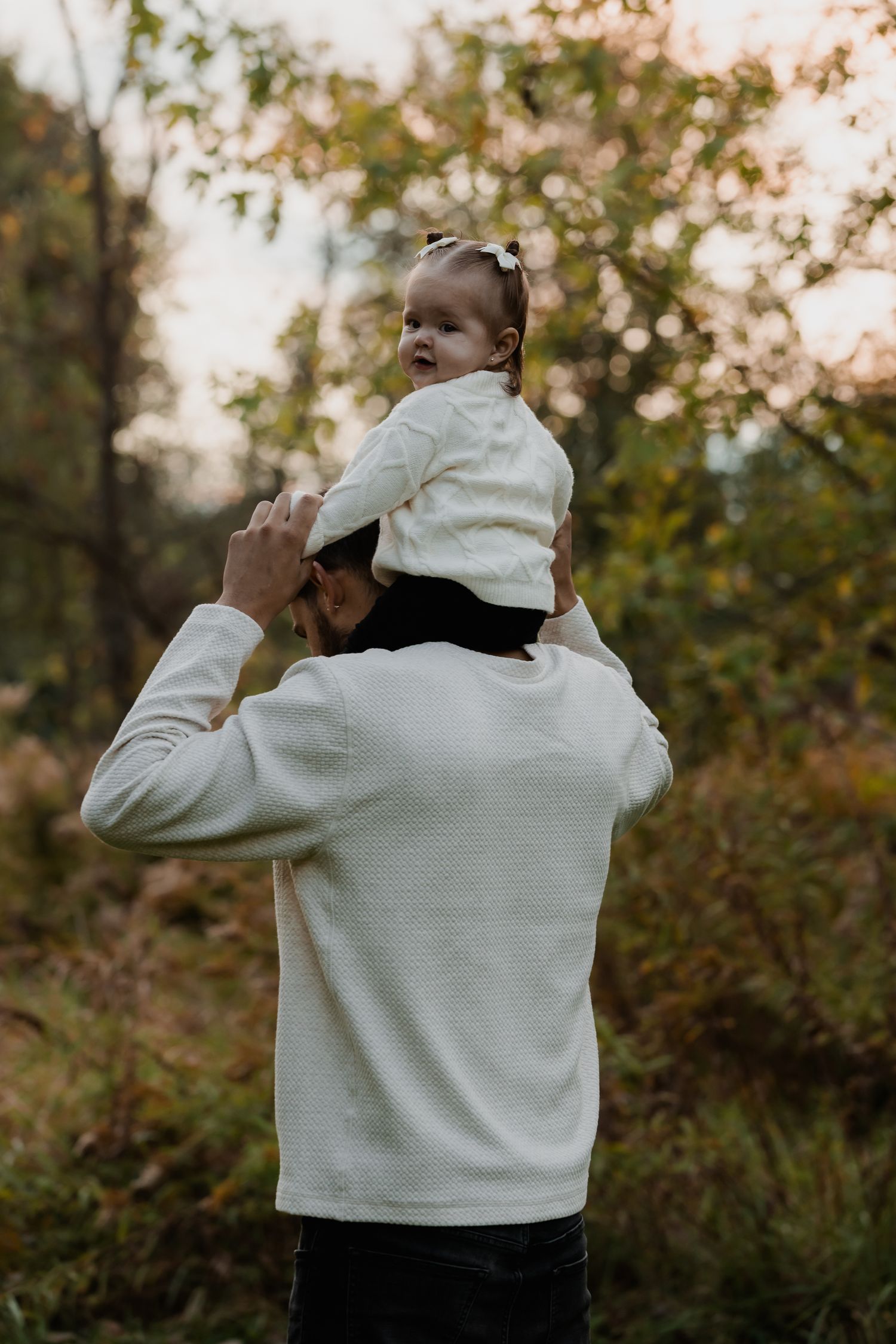Father holding baby on shoulders in autumn forest wearing matching white sweaters.
