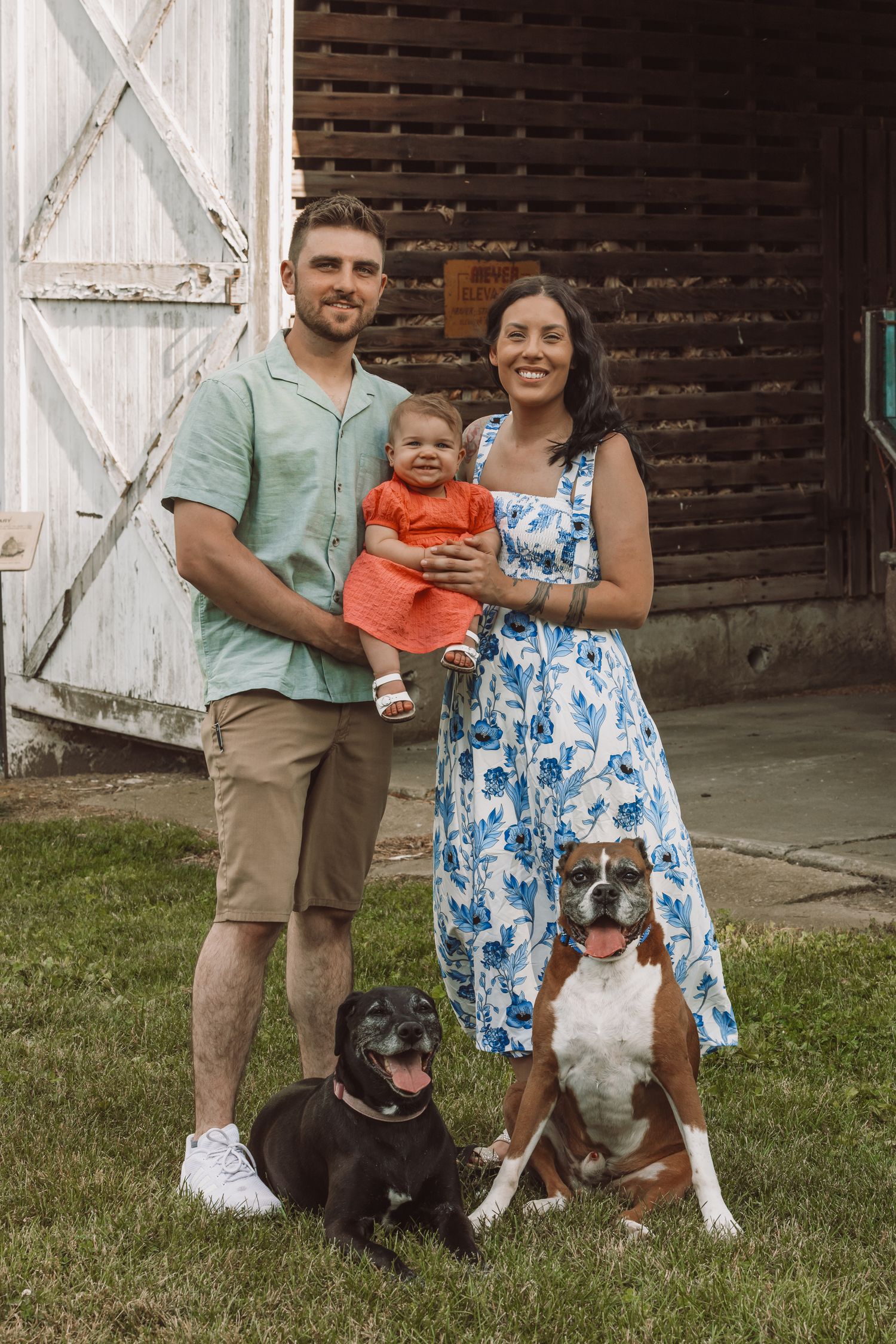 Family portrait with baby and two dogs in front of rustic barn.