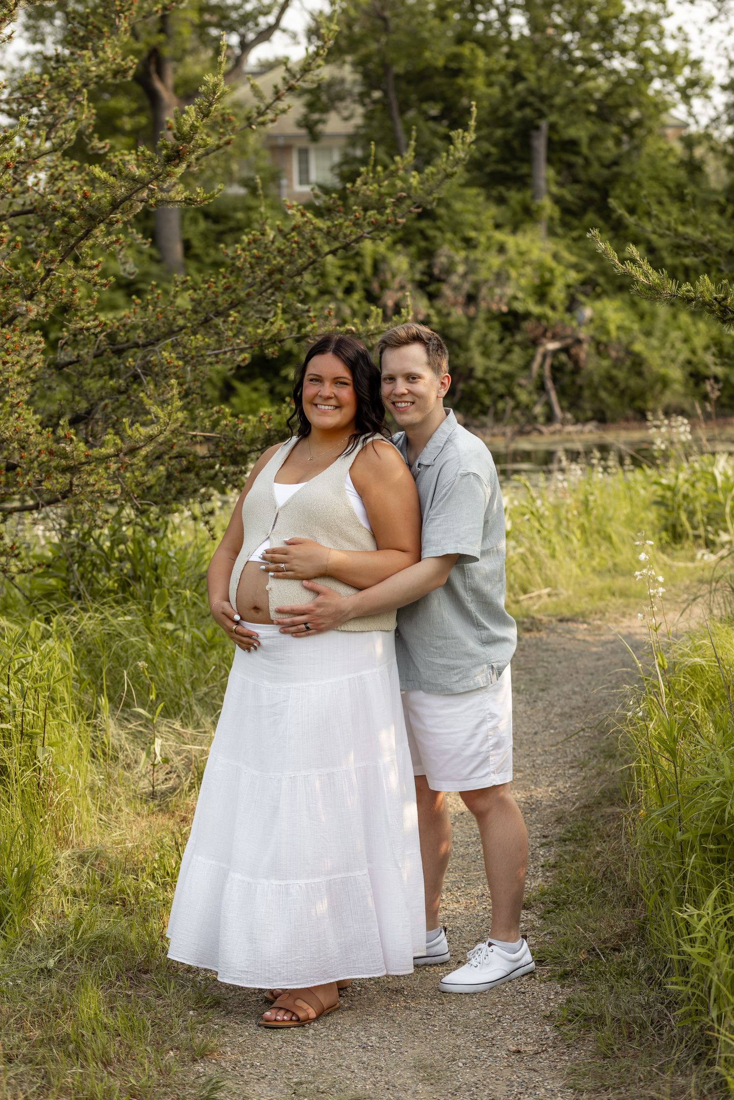 A couple poses outdoors on a path, the woman in a white dress cradling her pregnant belly, surrounded by lush greenery.