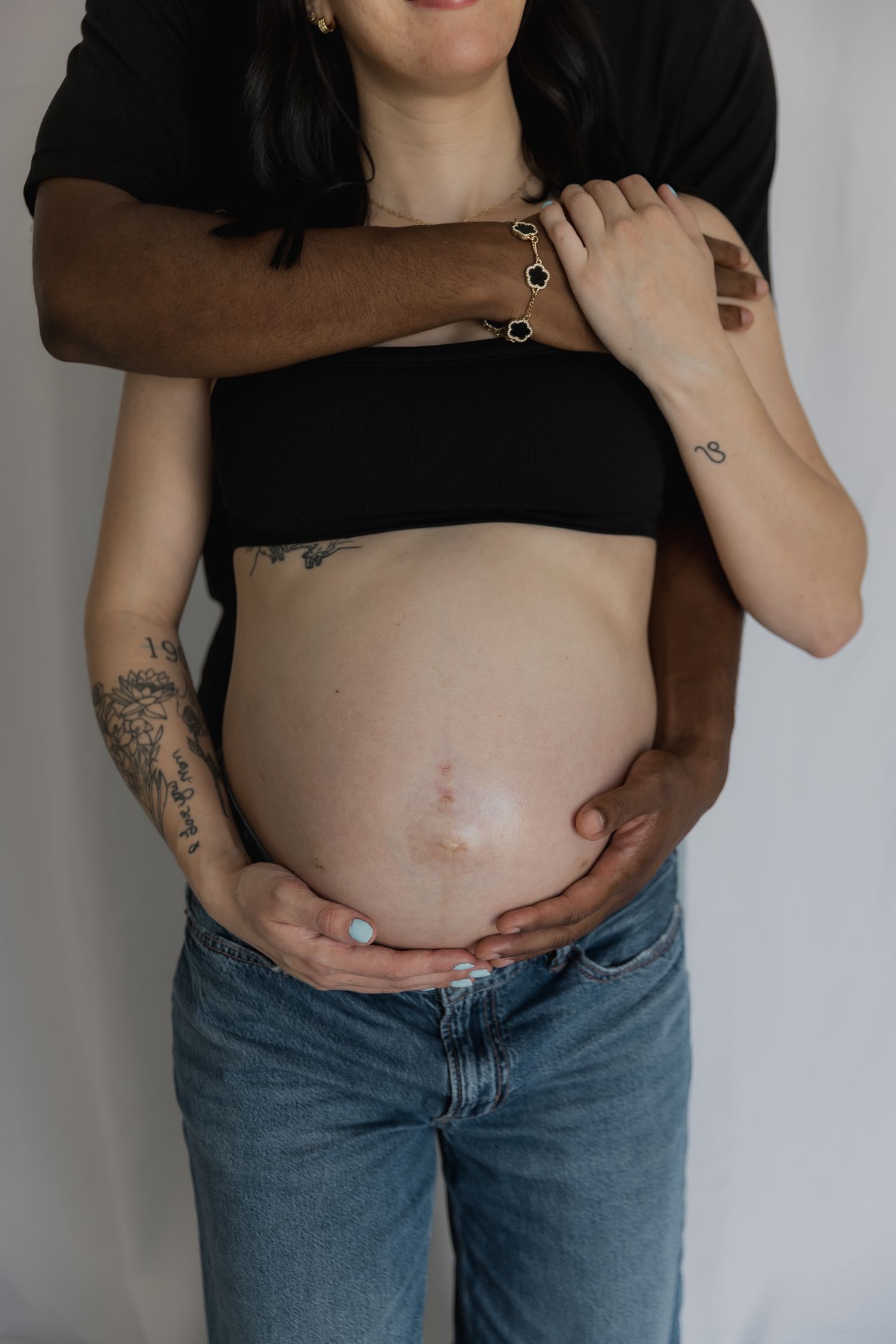 Pregnant woman with tattoos cradling her baby bump, partner embracing her from behind, maternity photo session.