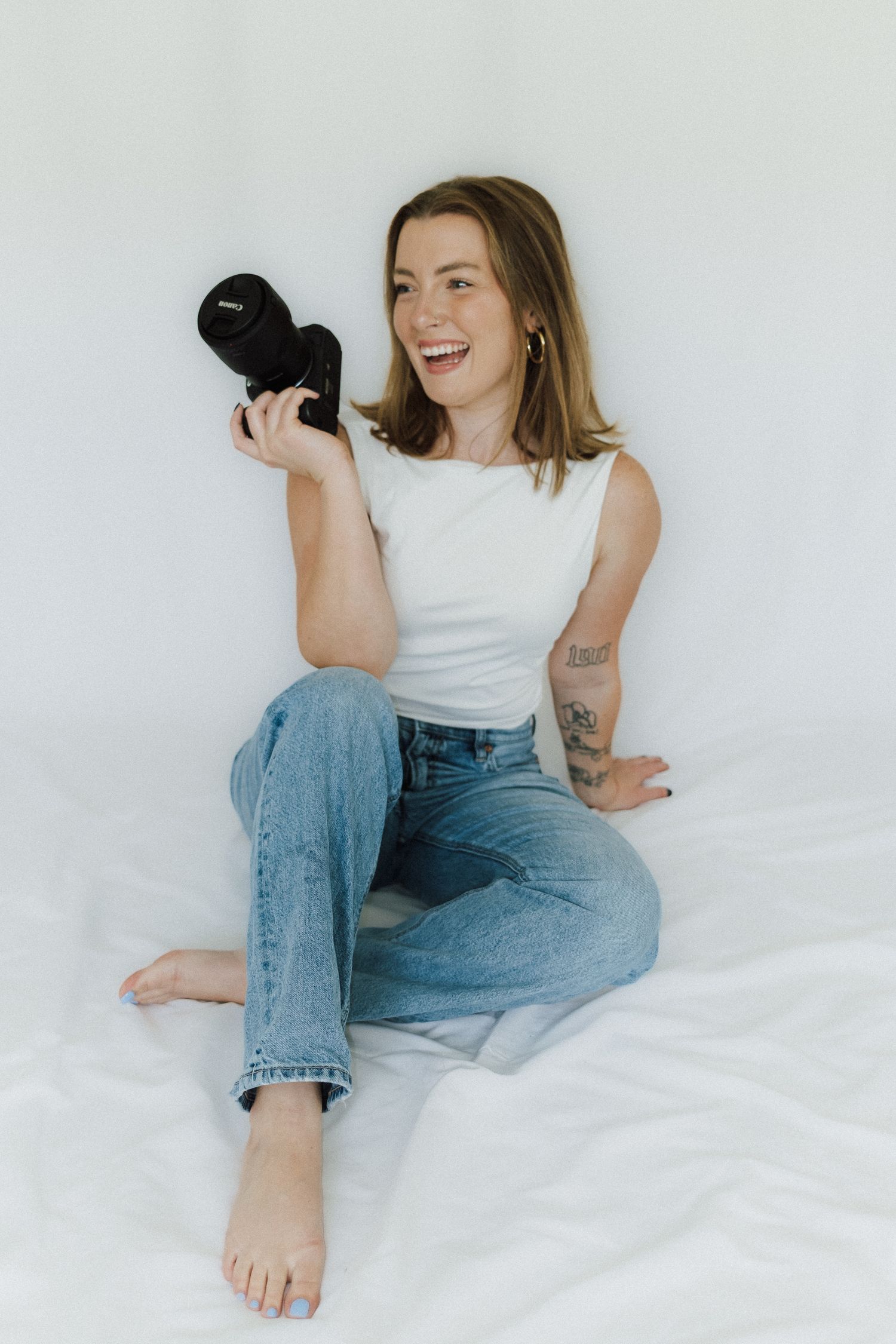 Smiling woman sitting on white bed holding a black camera, wearing white top and jeans with tattoos.