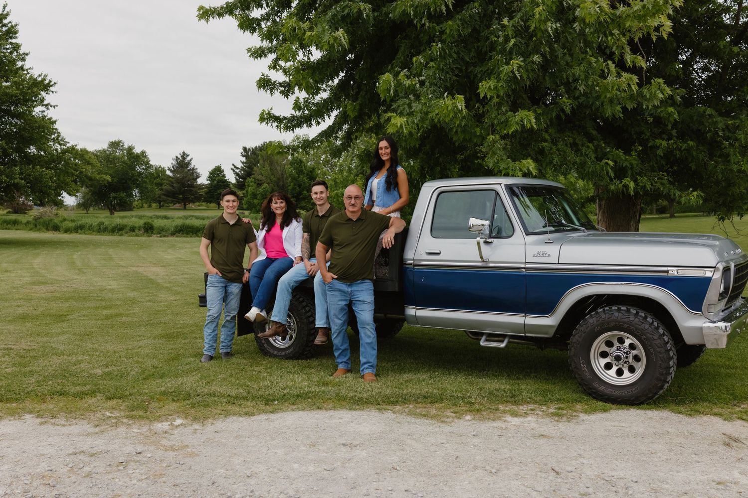 Family of five posing beside a classic blue and white Ford pickup truck on a green rural property.
