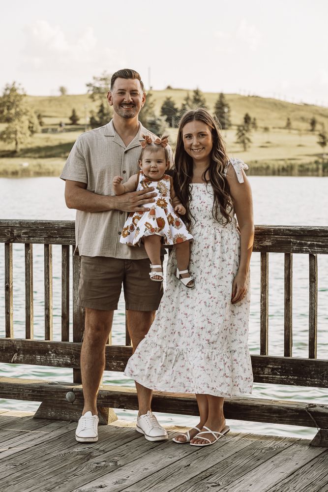 Happy family of three posing on a wooden dock by a lake, parents holding baby girl in floral outfit.