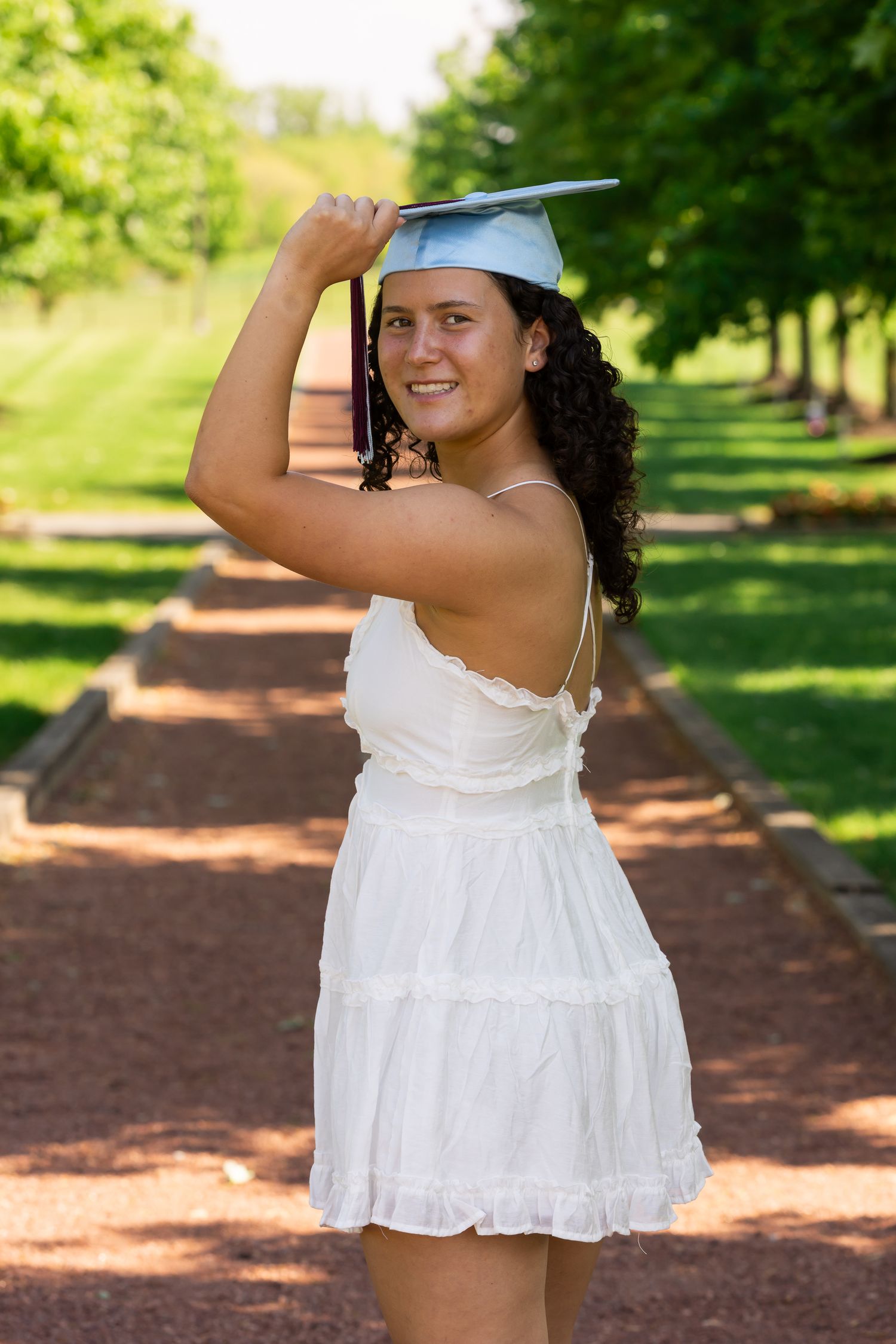 A young woman in a white dress holds a light blue graduation cap overhead, smiling on a tree-lined brick path.