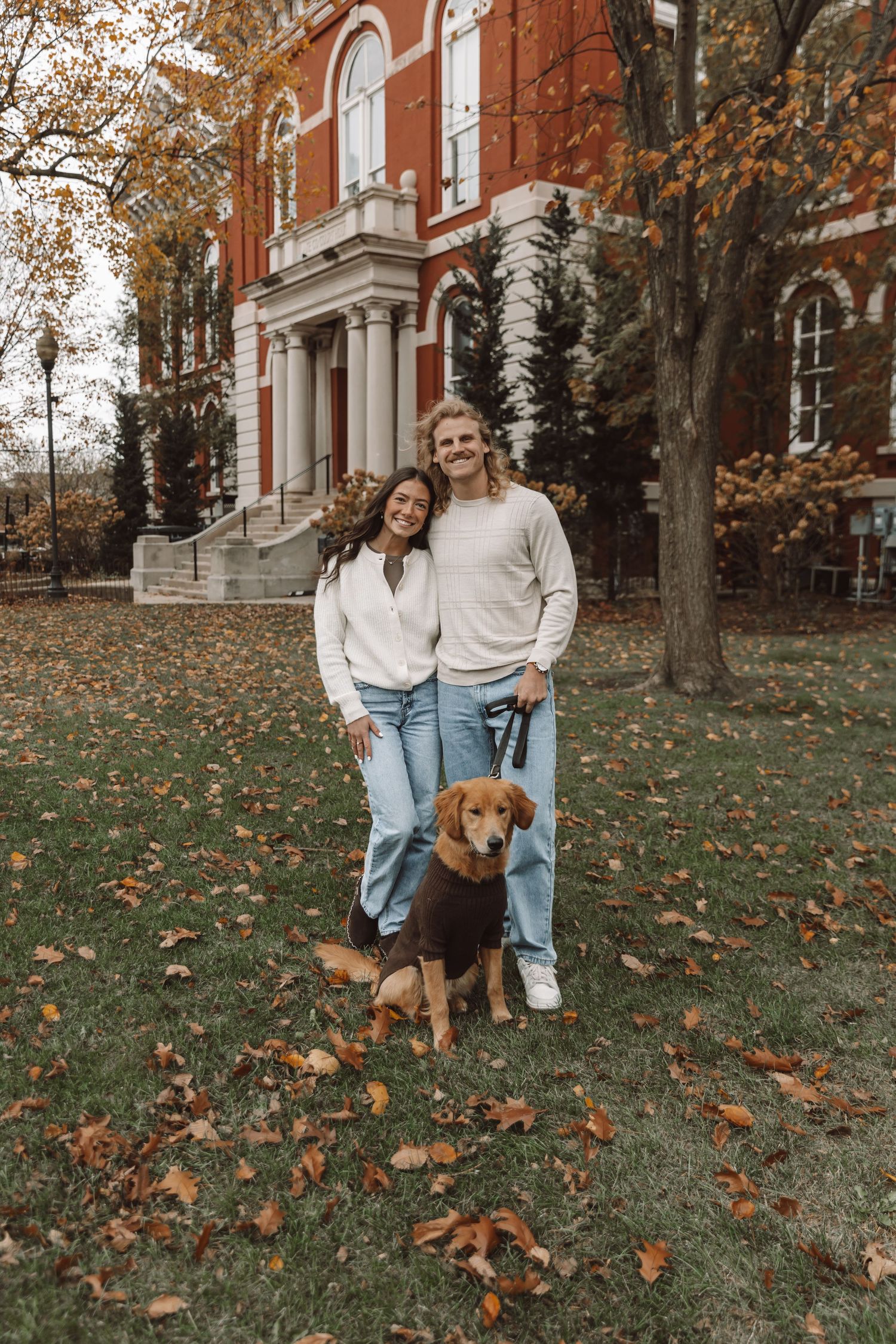 Couple and their dog posing on autumn lawn with red brick building in background.