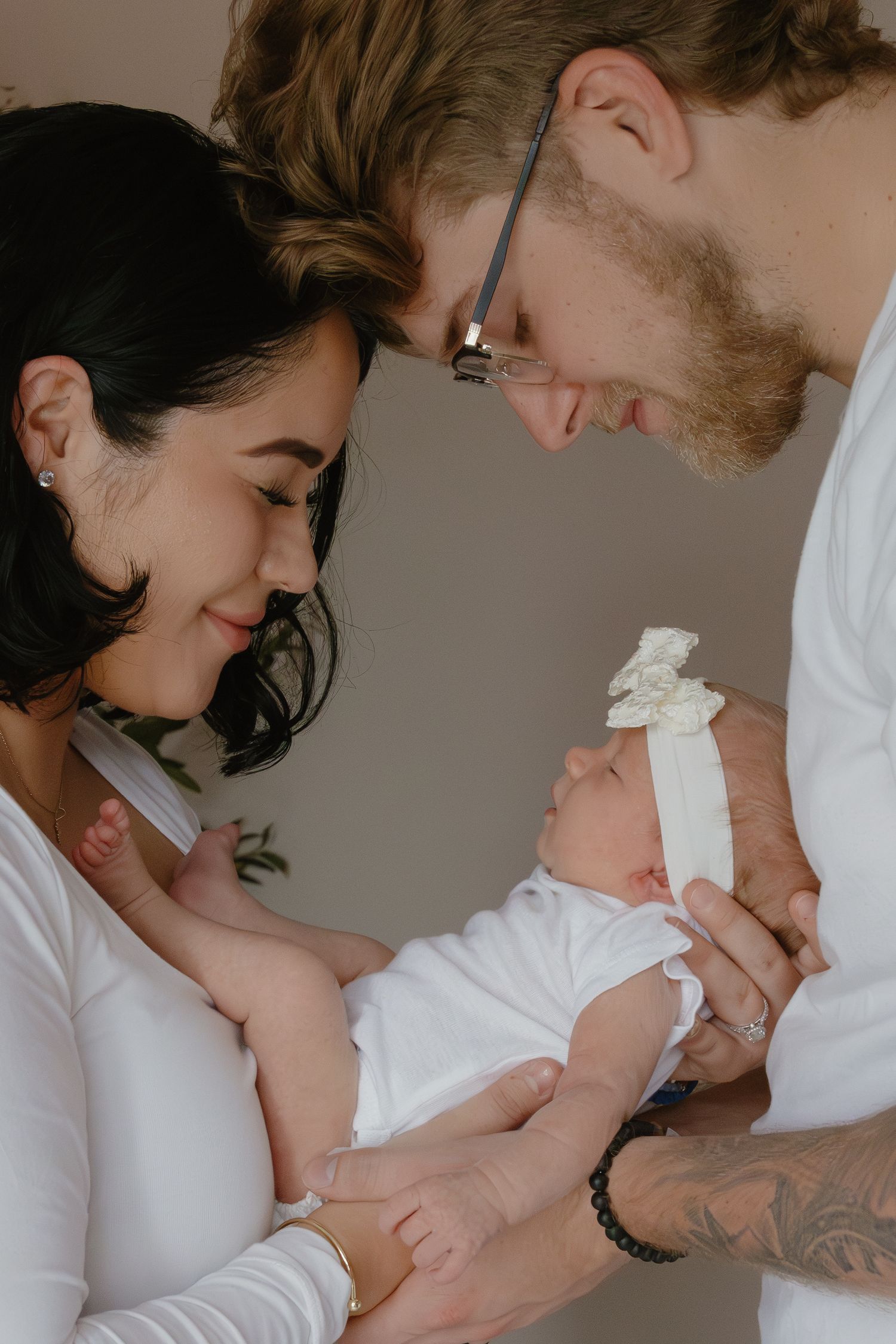 A couple in white tops lovingly gaze at their newborn baby dressed in white with a bow headband.