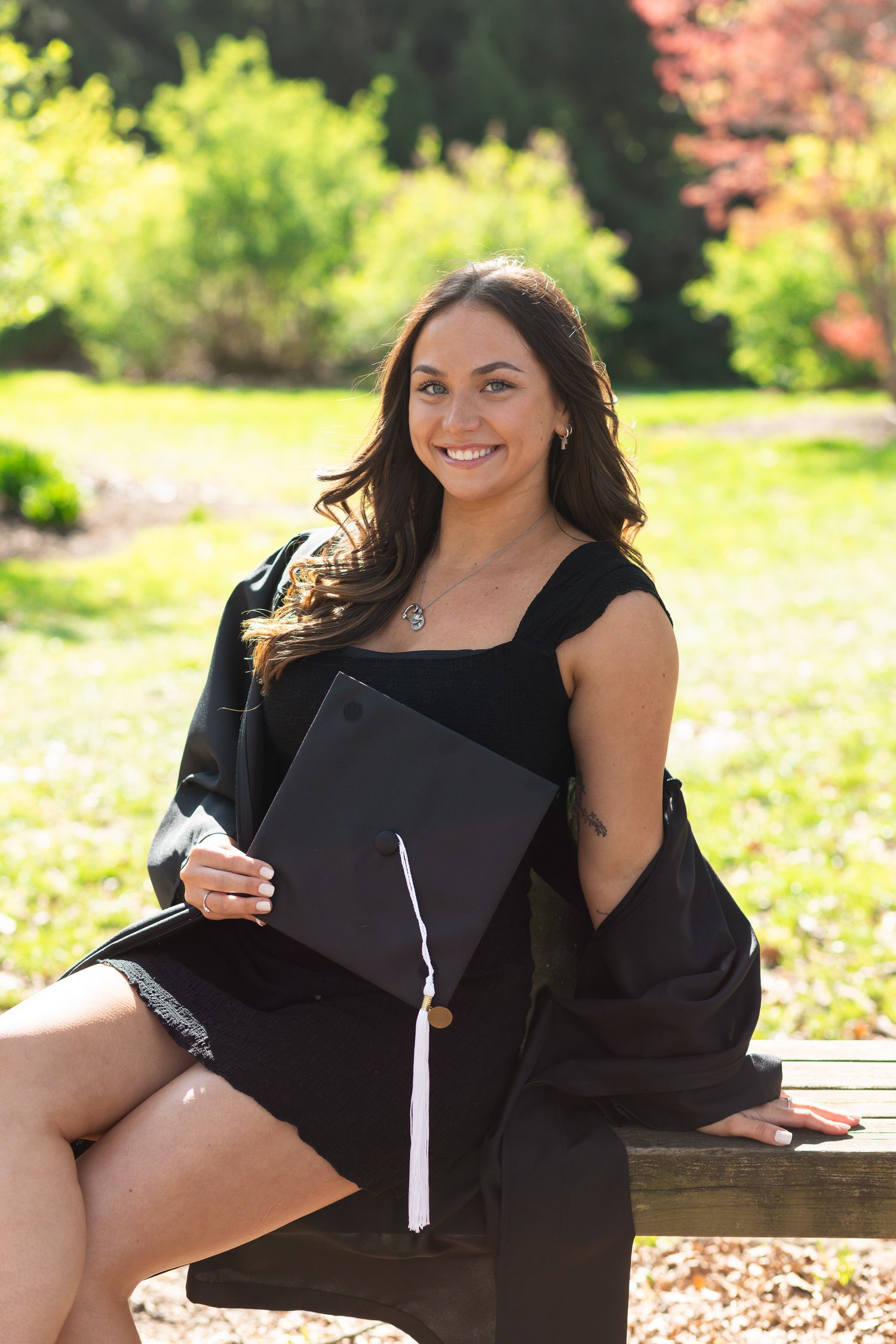 Smiling graduate in black cap and gown sitting outdoors on bench in lush green park setting.