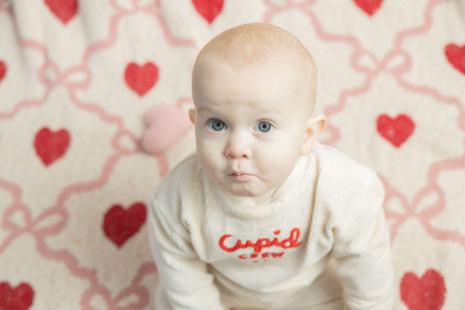 A baby wearing a white sweater sits against a background decorated with red hearts and pink swirls.