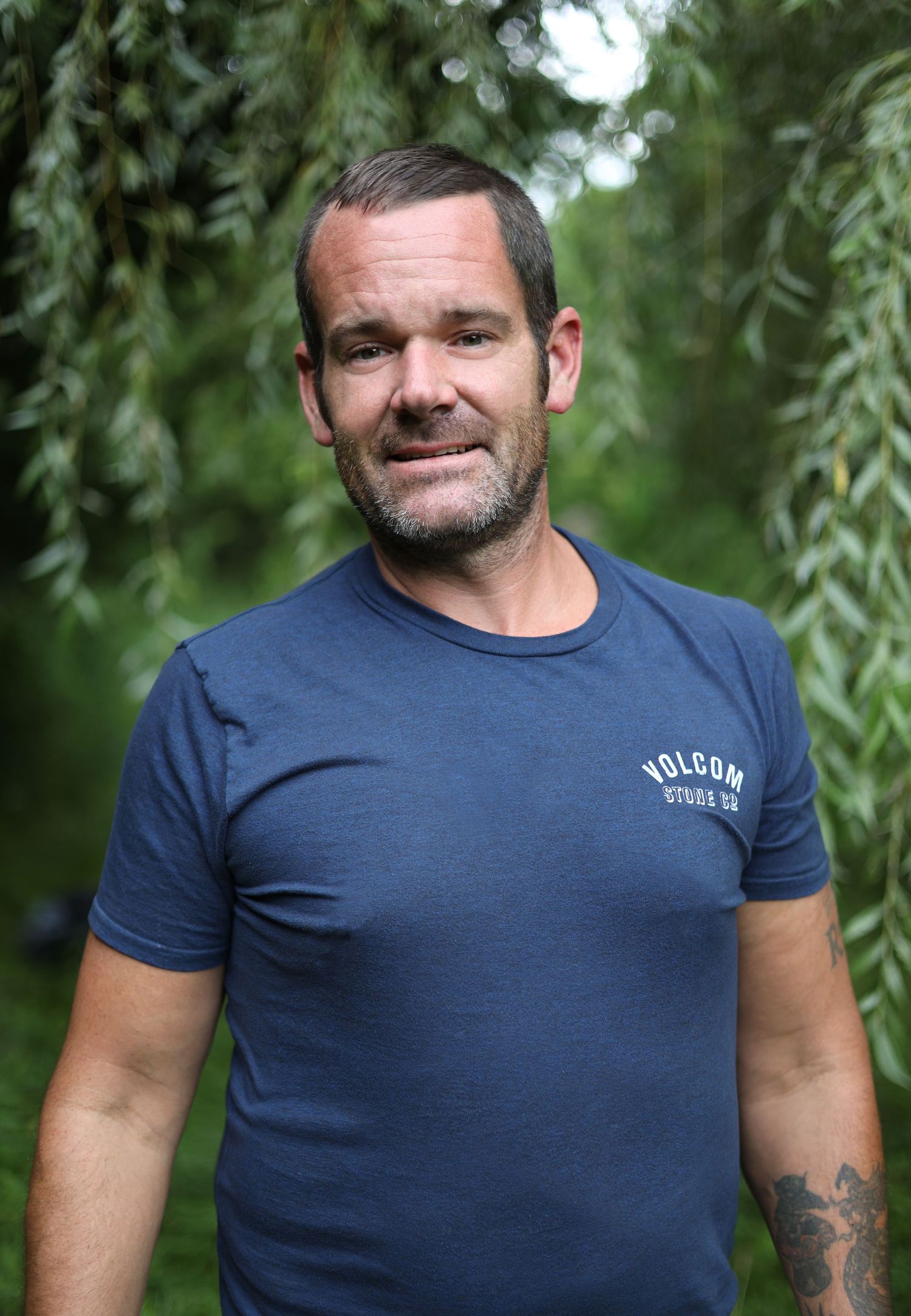 Person wearing a navy blue t-shirt with a small logo stands in front of green foliage in an outdoor setting.