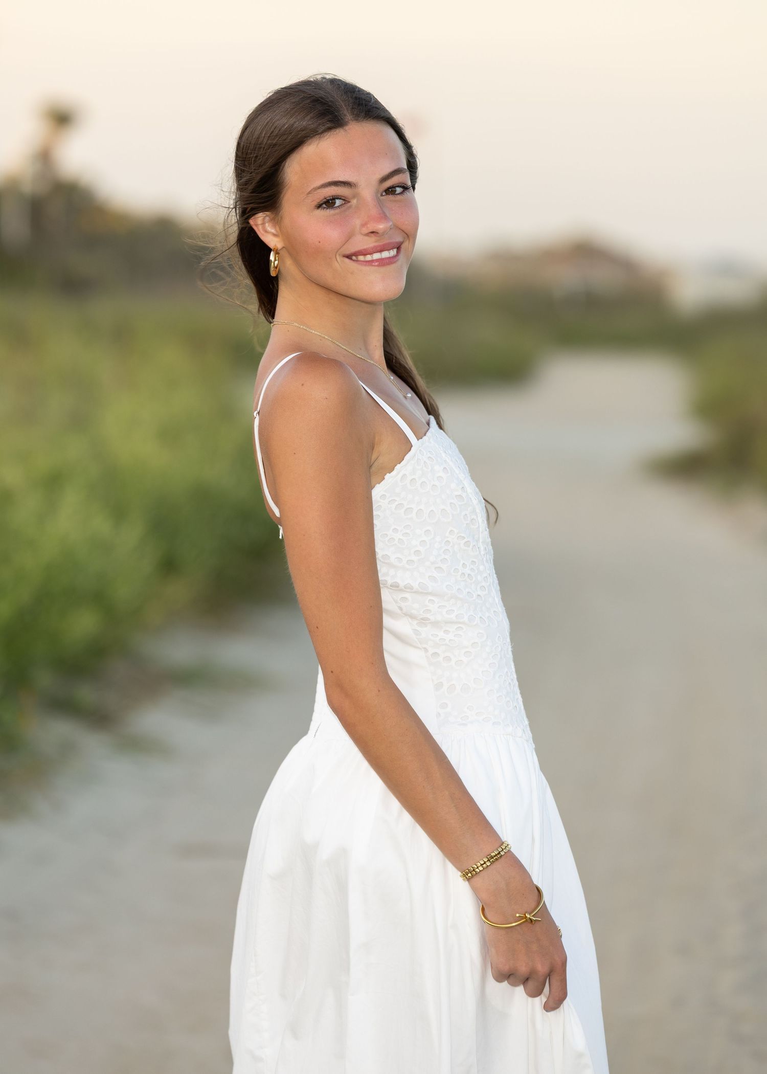 High school senior girl looking over her shoulder at the camera while smiling with a sandy road and dunes behind her.