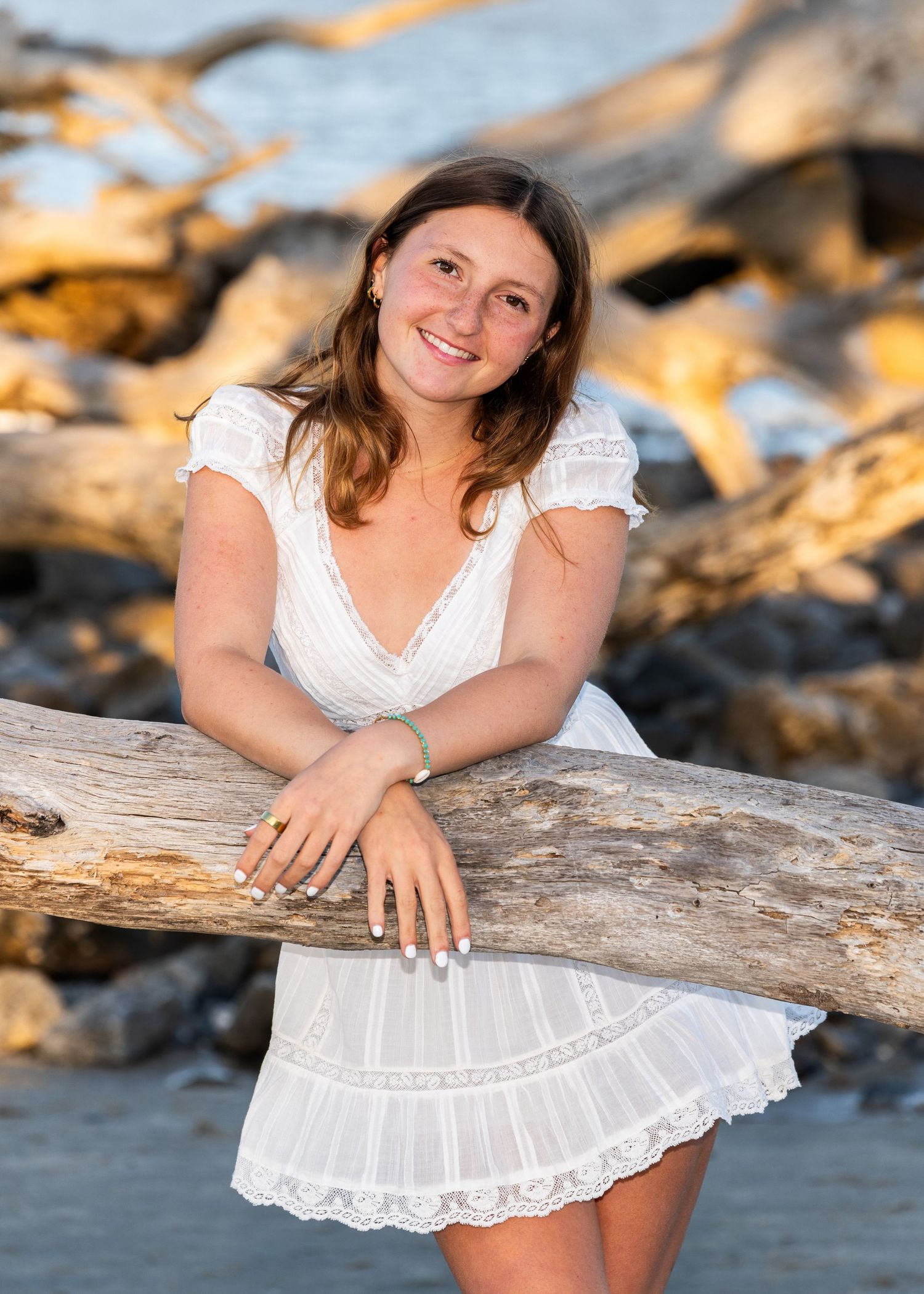 A high school girl senior leaning on a tree at Driftwood Beach, with her hands draped over the tree and smiling at the camera.