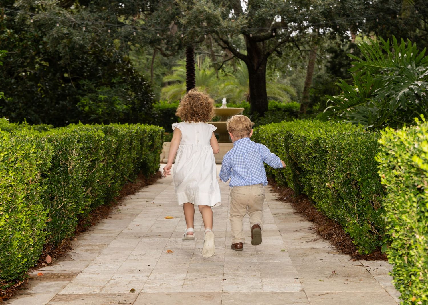 A small girl and boy are running away from the camera down a stone pathway surrounded by green bushes.