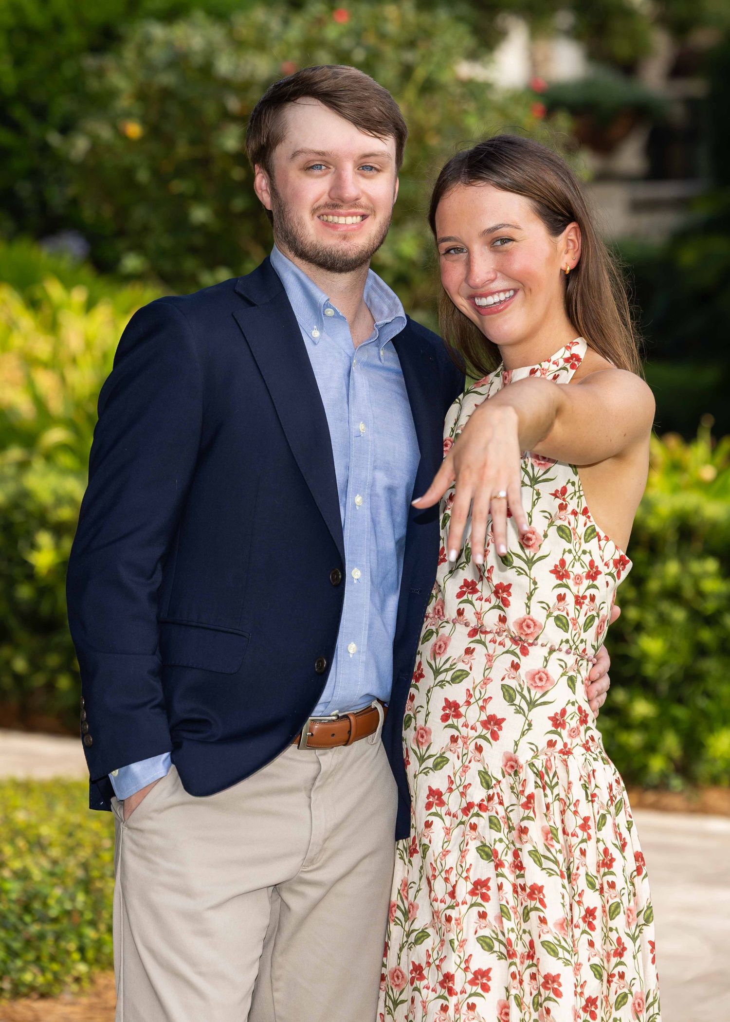 A couple smiling at the camera. She is holding her hand out, showing off her new engagement ring. He just proposed