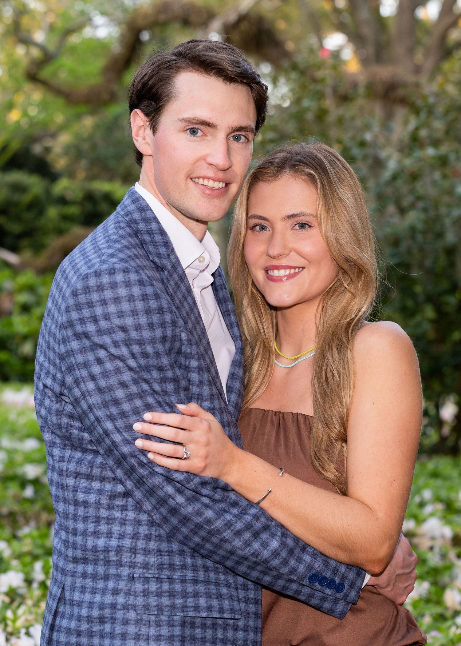 A couple smiling at the camera. She has her hand on his arm so you can see her new engagement ring. He just proposed.