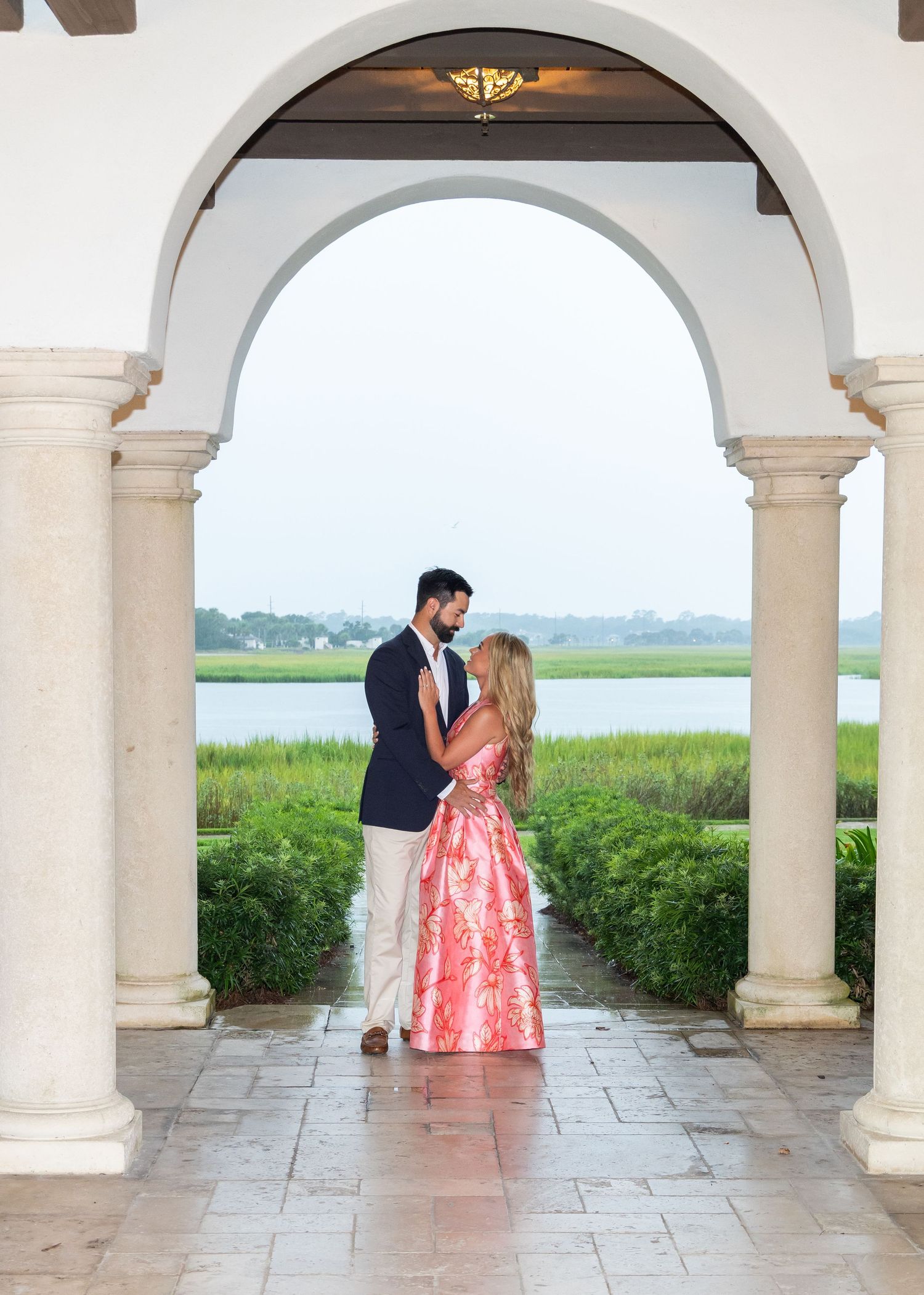 A recently engaged couple standing in the distance under an arch way with the marsh in the background.