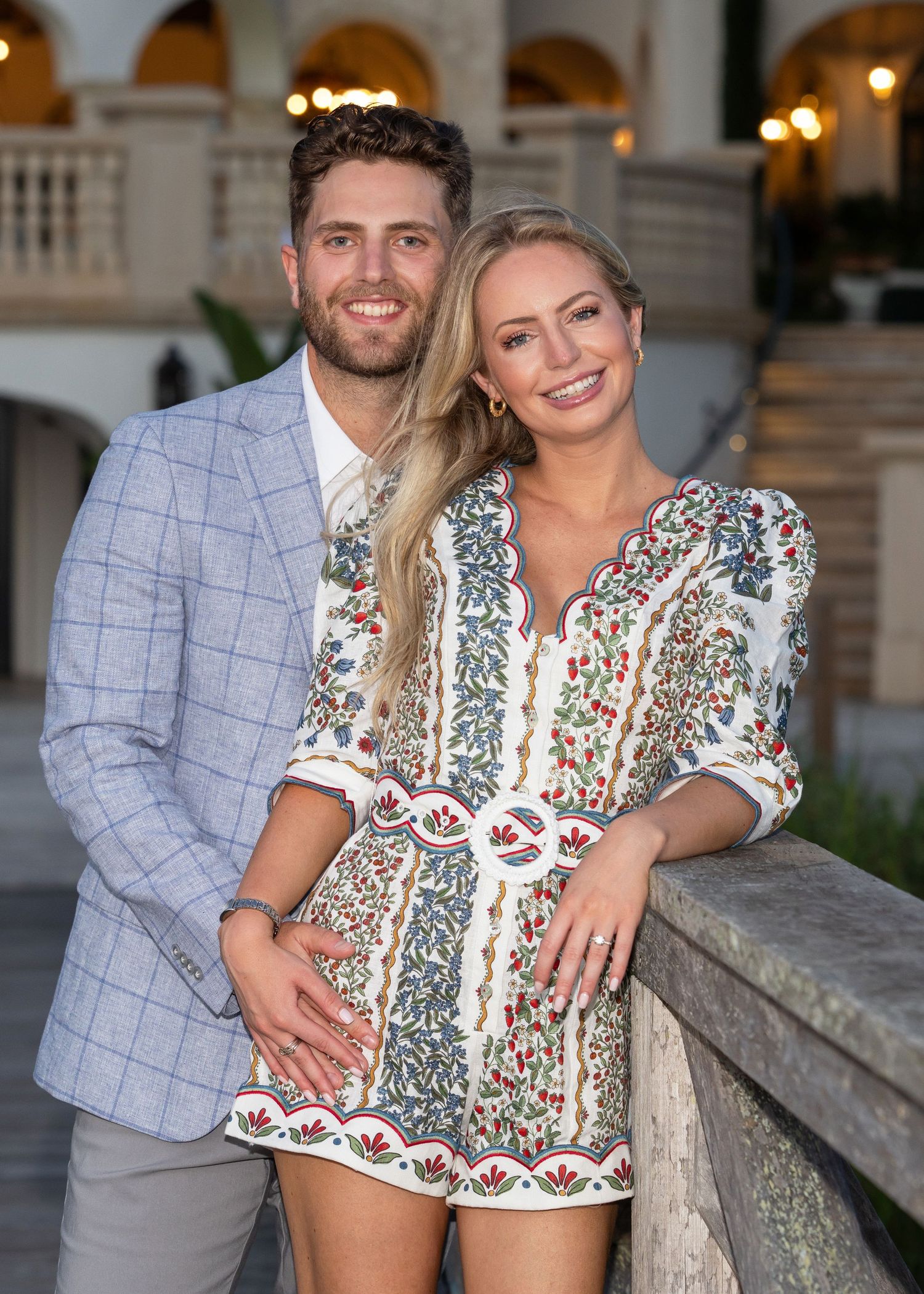Couple standing on a dock. She is standing in front with her arm resting on the railing as her hand dangles showing off her engagement ring.