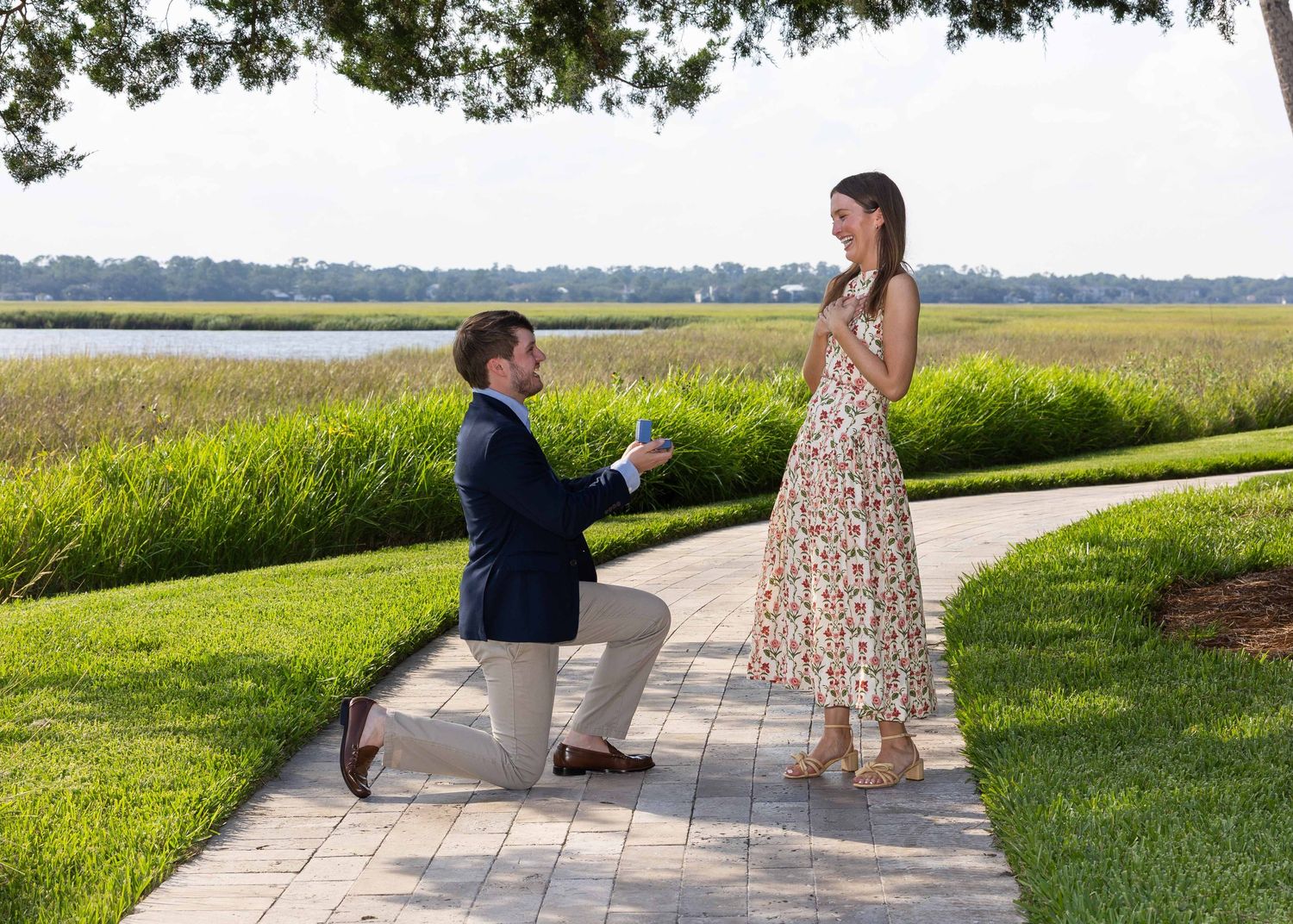 A proposal where he is on one knee with ring box in hand and she is standing with her hands on her chest looking pleased and surprised.