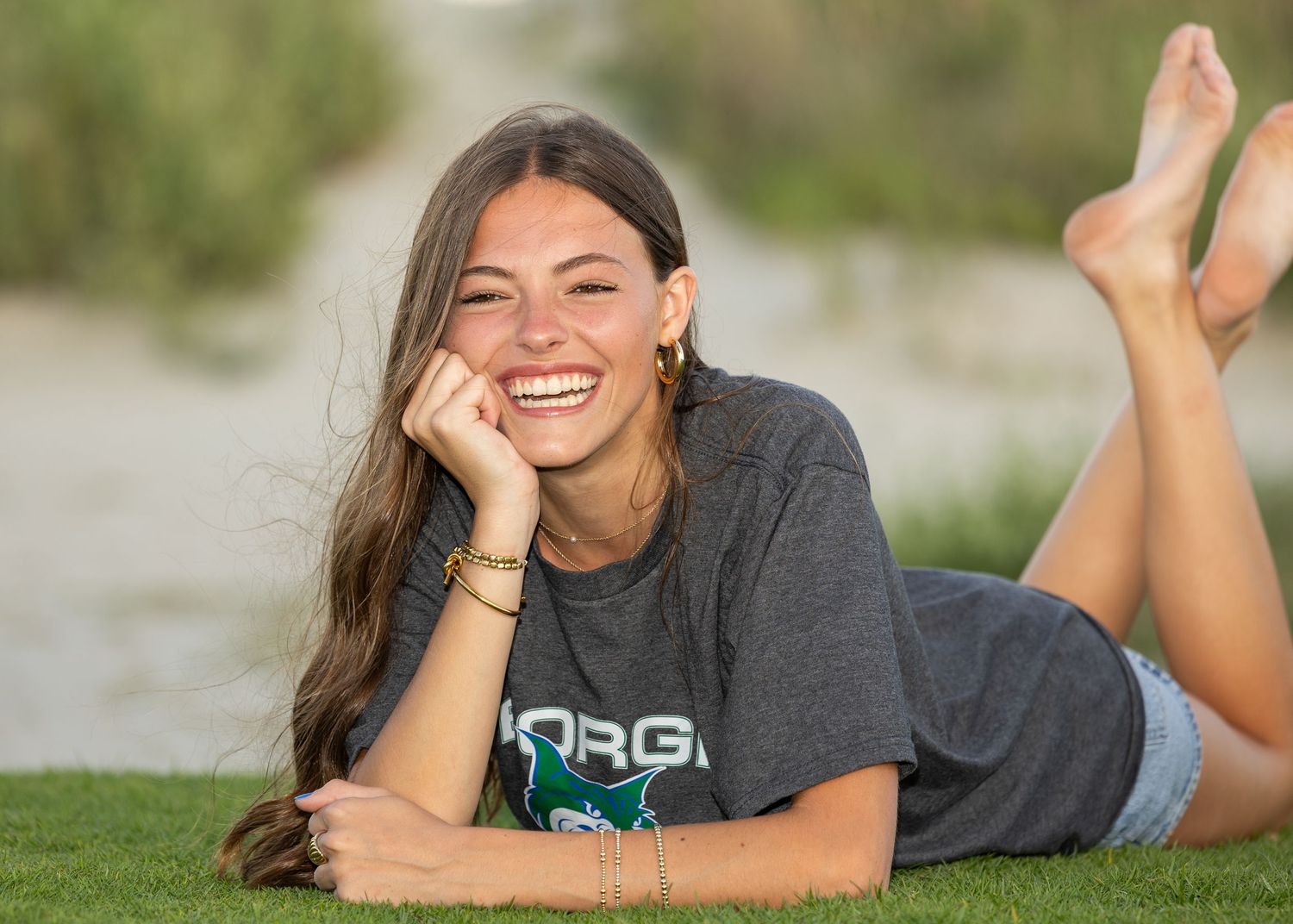 High school senior girl laying on her stomach with her feet kicked up, laughing at the camera while wearing her college t-shirt.