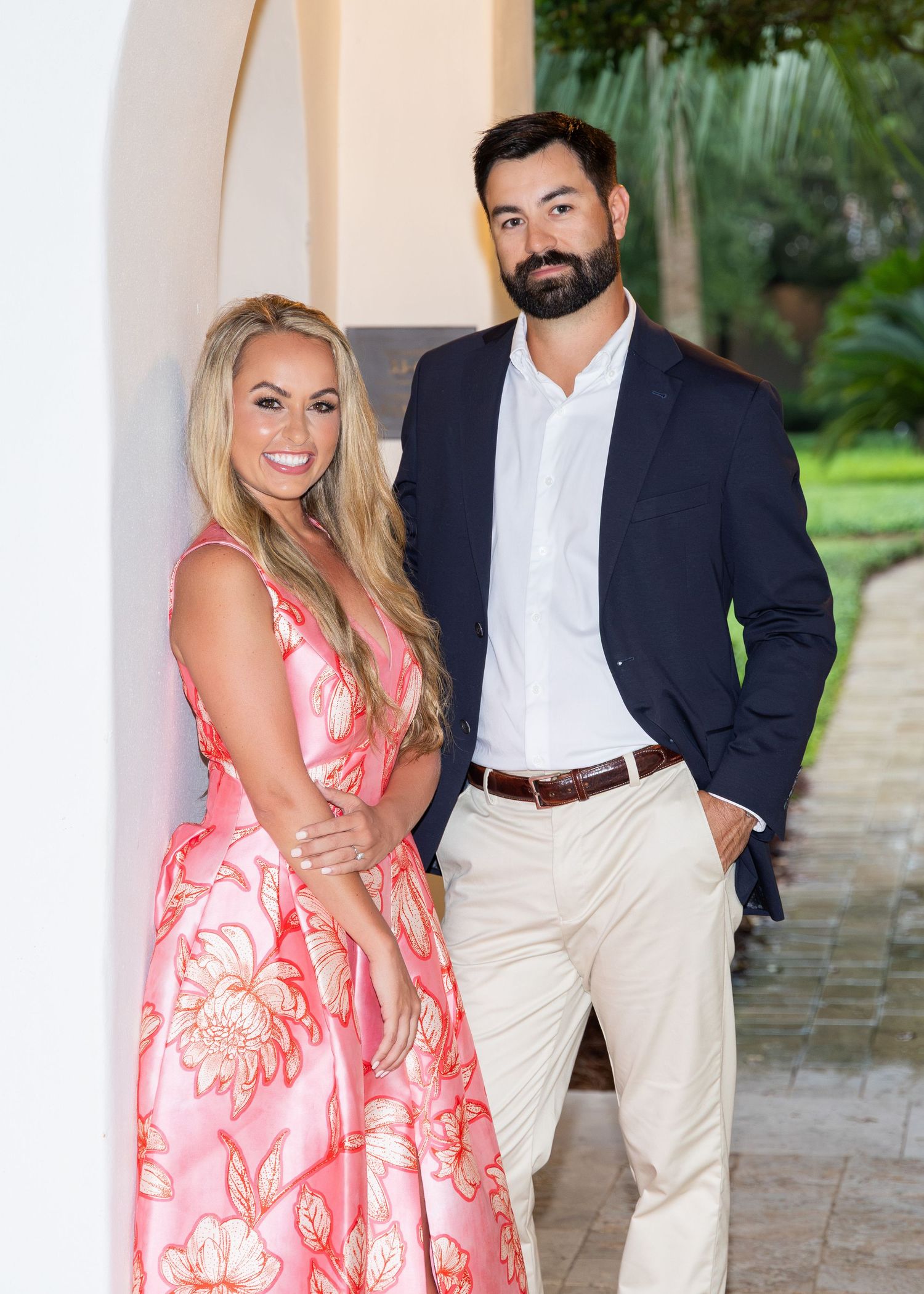 A recently engaged couple standing under an archway. He is behind her as she leans against the wall and both are smiling at the camera.