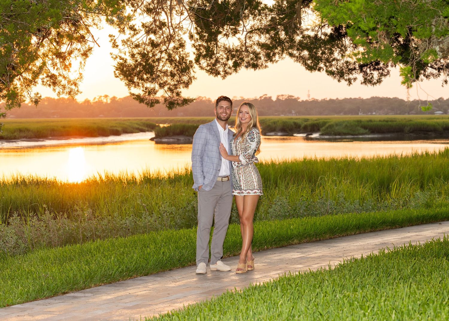 A couple standing on a stone pathway under trees with the sunsetting behind them. Her hand is on his chest showing off her engagement ring.