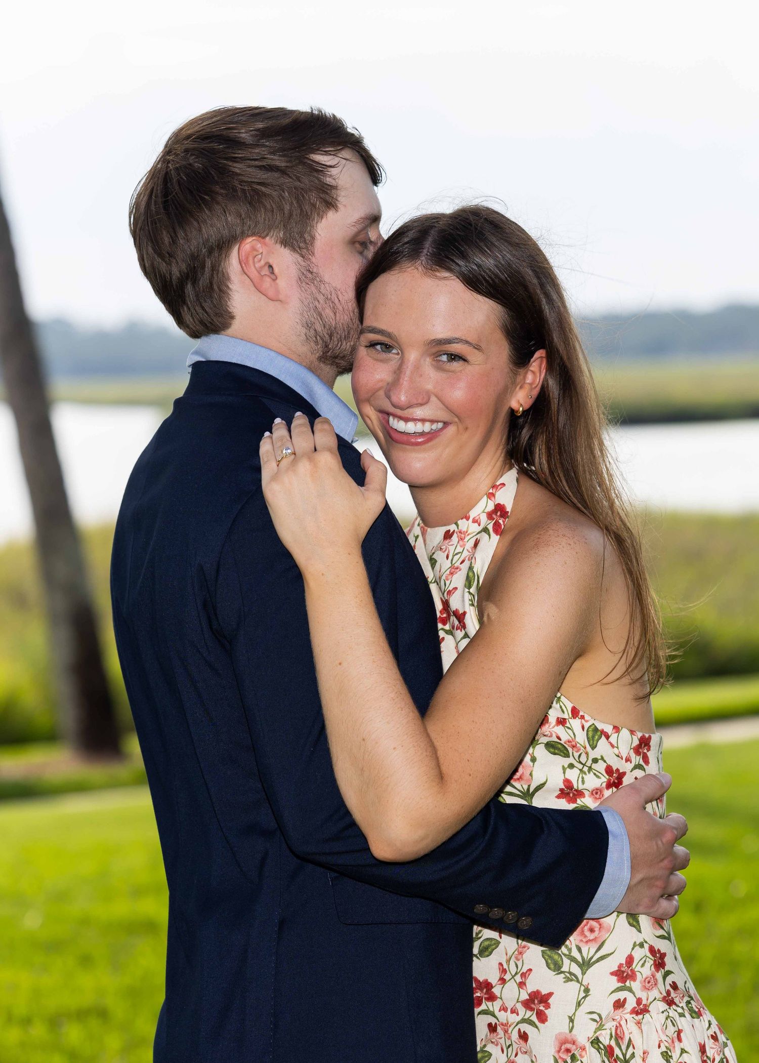 A couple hugging. She is laughing at the camera and he is looking away. Her hand is resting on his shoulder showing of her engagement ring.