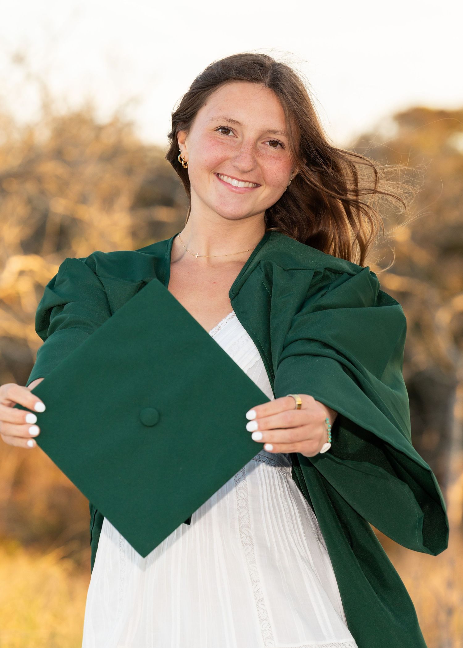 A high school senior girl wearing her graduation gown while holding her cap out to the camera and smiling at the beach.