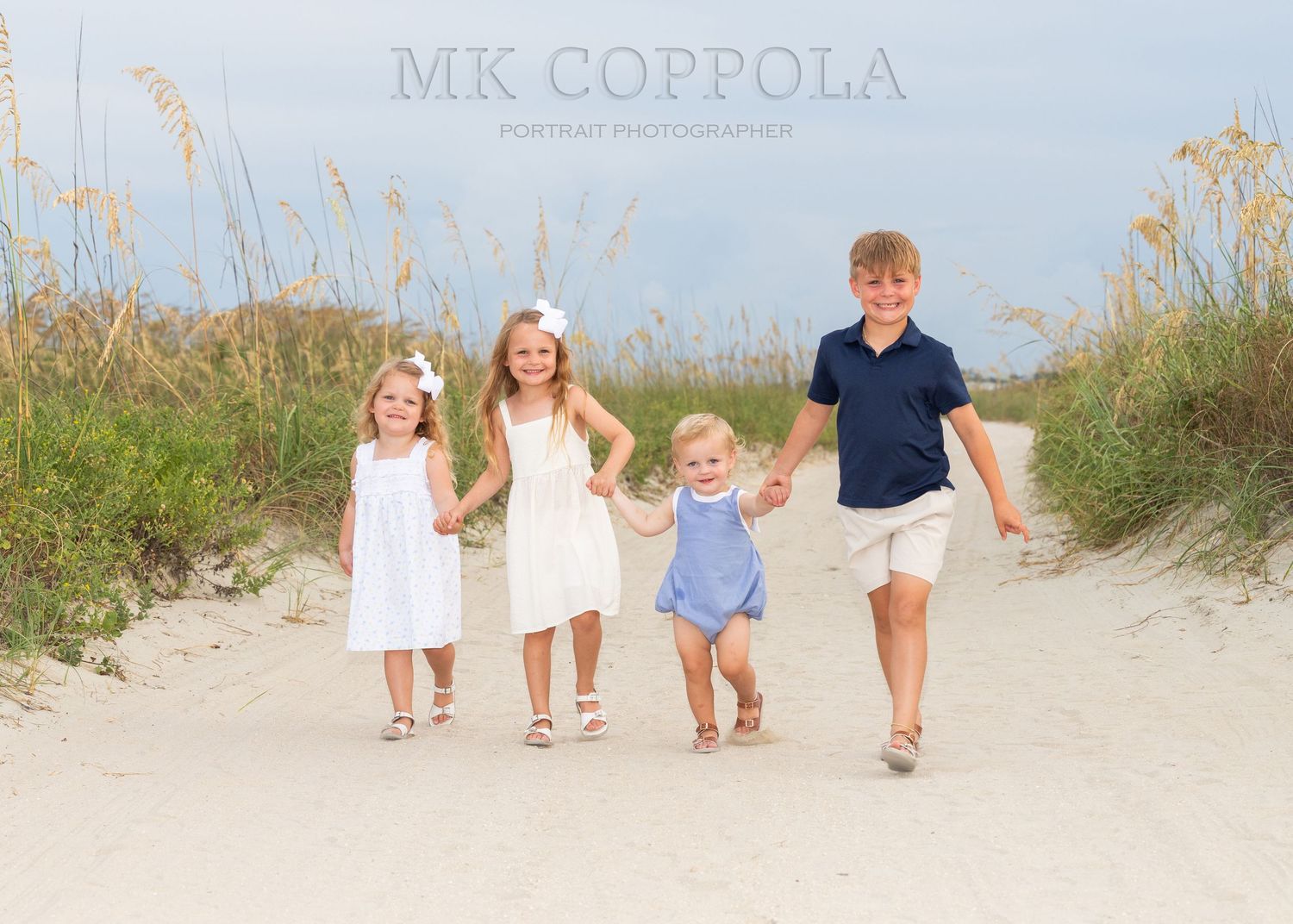 4 children walking on a sandy road, smiling and holding hands while looking at the camera.